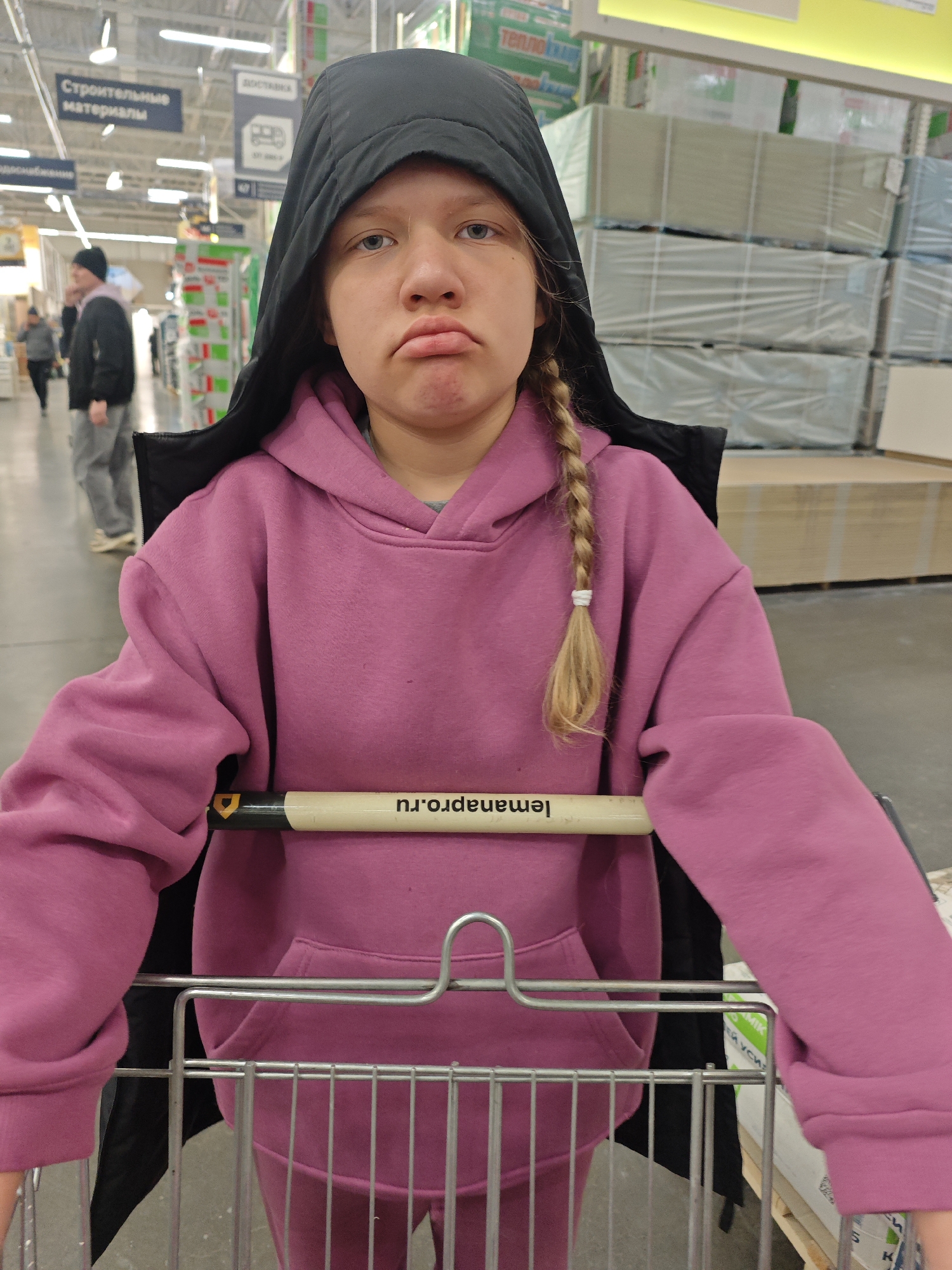 A young person stands indoors in a retail environment, possibly a hardware or home improvement store, pushing a shopping cart. The individual is wearing a pink hoodie and a black hooded jacket, with blonde hair braided on one side. Their facial expression appears sullen.
The shopping cart has the word "lemana.pro.ru" printed on the handle, which is readable when mirrored. In the background, shelving units are stocked with construction materials. A sign in Russian visible reads "Строительные материалы" (construction materials). Other people are walking in the background.
Based on the "lemana.pro.ru" URL and the Russian signage, the location is likely a branch of the Leroy Merlin chain in Russia, and potentially the store located in Izhevsk.