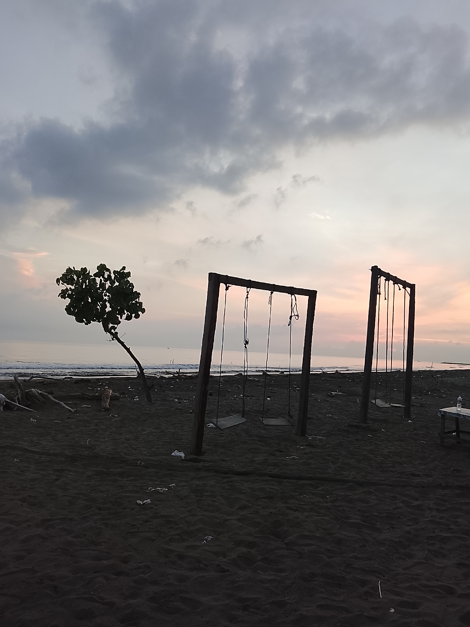 The image depicts a sandy beach at dusk in Reudeuep, Indonesia, with the ocean in the background. Two wooden swing sets with seats are visible on the dark sand. To the left, a solitary tree with a dark canopy leans towards the sea. Debris, including driftwood, is scattered across the beach. The sky is cloudy, with hues of pink and gray, suggesting a sunset or sunrise. The water is calm and reflects the muted colors of the sky. A small table with a water bottle is present on the right side of the frame.