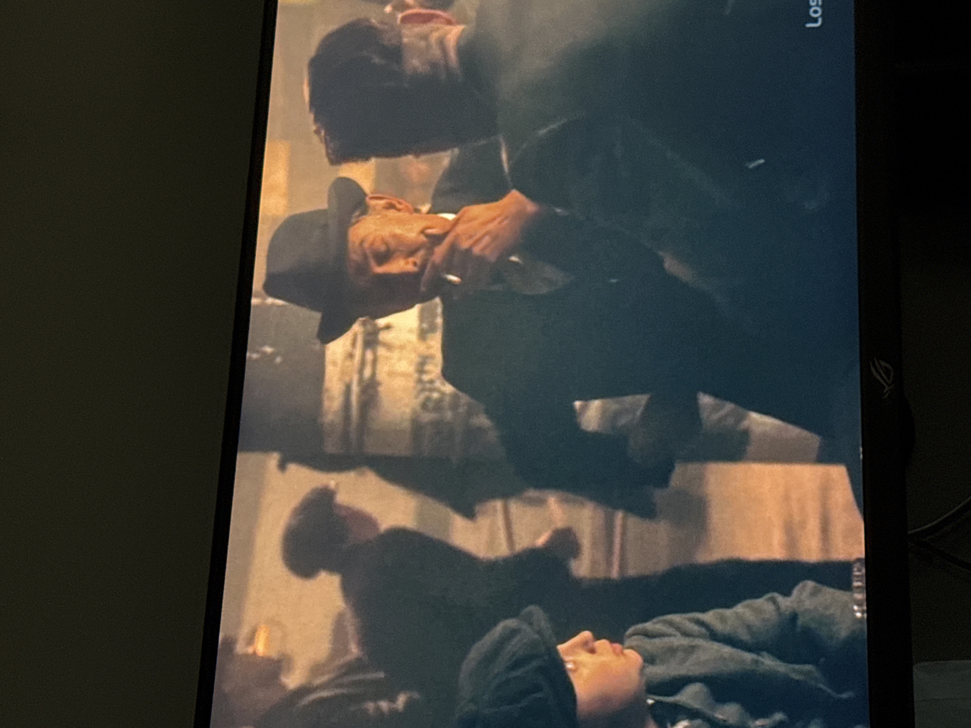 A man in a dark suit and fedora holds a cigarette and looks down at another man lying prone, with another man lying in the background; likely in a dimly lit indoor environment.