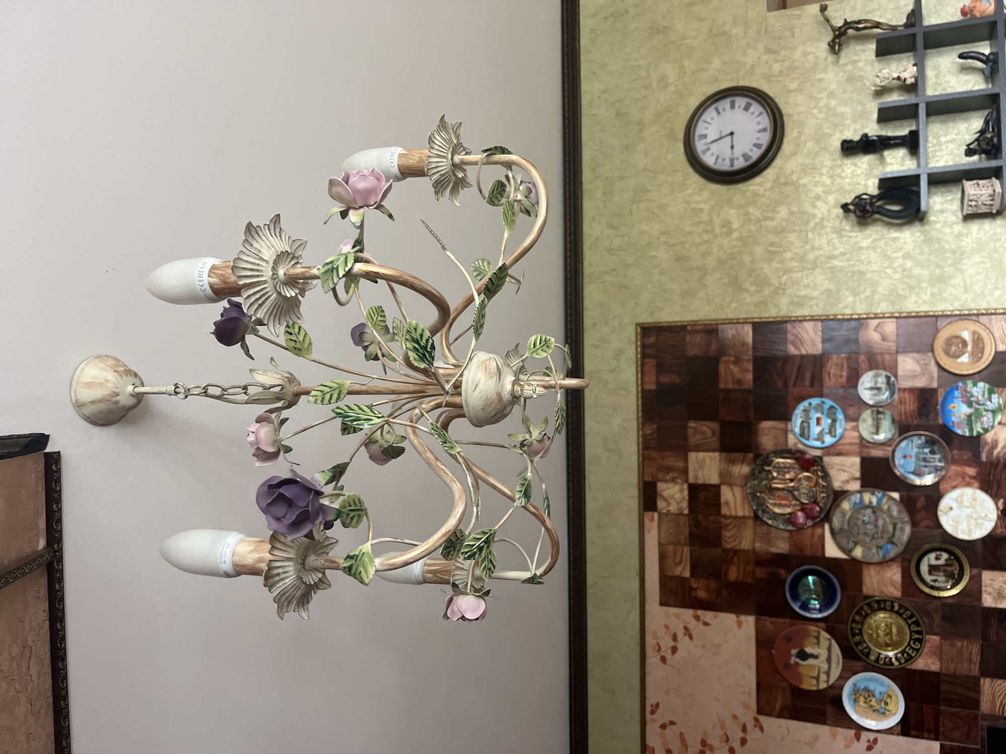 This is a medium-angle, indoor shot of a cream-colored chandelier adorned with artificial flowers and leaves, hanging from a white ceiling. The chandelier has multiple arms, each ending in a candle-shaped light bulb fixture, some of which hold white, elongated bulbs with visible text. The background features a textured, light green wall with a dark wood frame running horizontally across it. To the left of the frame is a large, decorative piece of art composed of a checkered pattern of dark wood squares and rectangles, interspersed with decorative plates. Below the chandelier, a portion of a wall clock with Roman numerals and black hands is visible. Further to the right, a dark gray shelf holds several small decorative figurines.