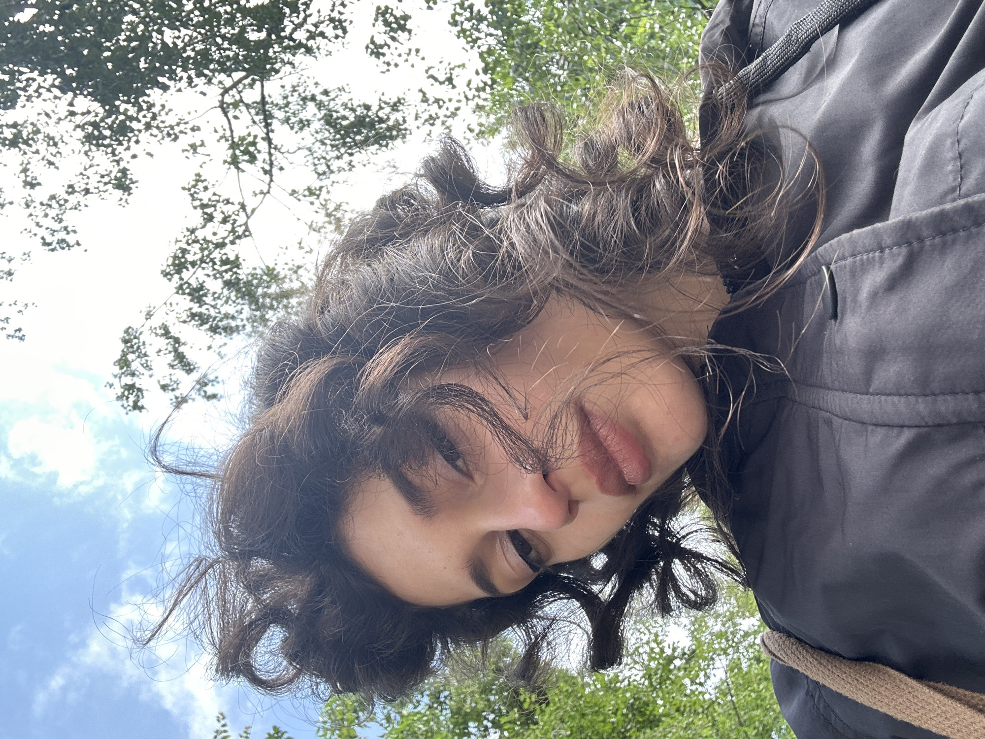 A low-angle, close-up, outdoor selfie shows a young woman with dark, curly hair against a bright sky with scattered clouds and blurred green foliage. She is wearing a black jacket and a beige strap is visible over her shoulder. The lighting suggests it is daytime. No specific location in Sofia, Bulgaria is identifiable from the image.