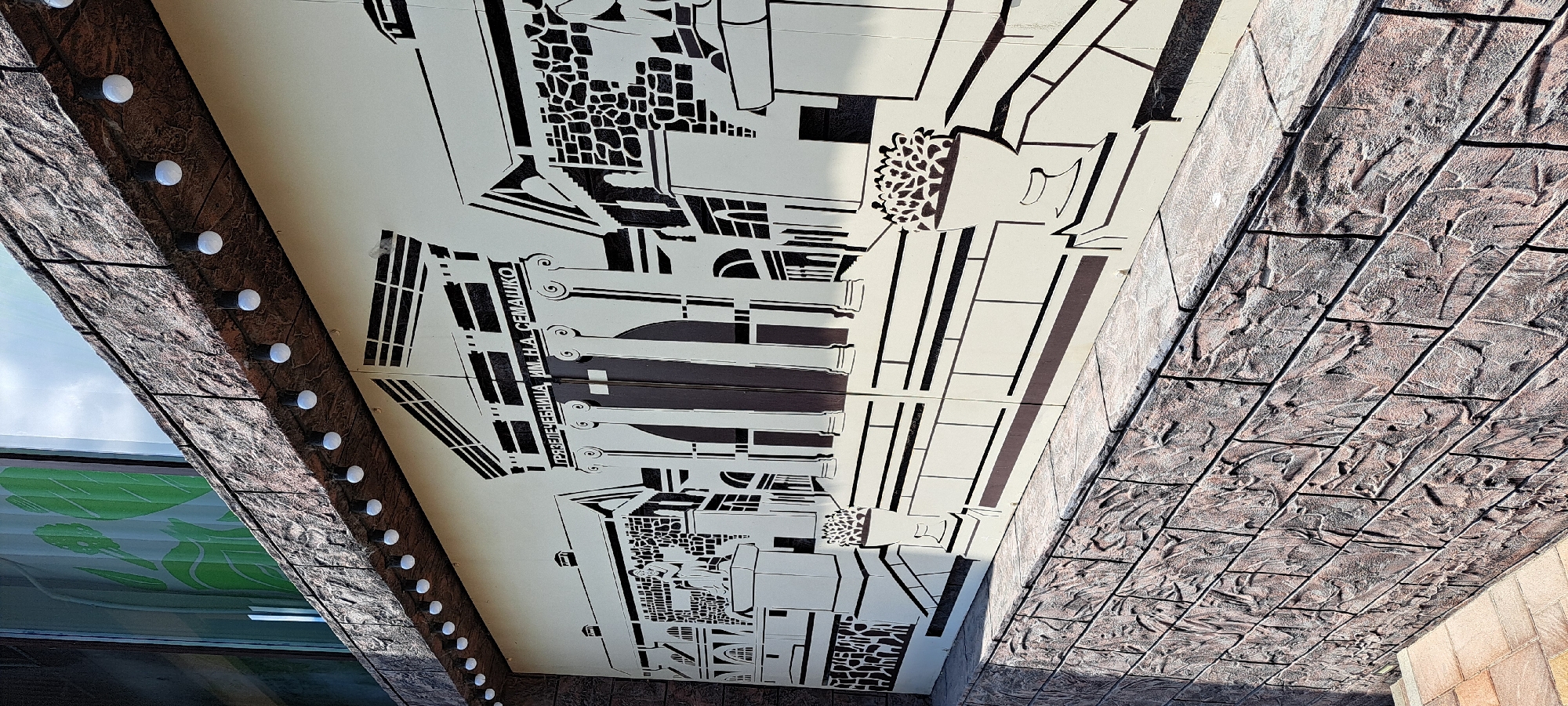 The image shows a mural on the facade of a building, depicting a stylized architectural scene with classical elements. The mural is rendered in black and white and features columns, a pediment, and what appears to be a statue. Text in Cyrillic script is visible above the central columns, reading "ГРЯЗЕЛЕЧЕБНИЦА ИМ. Н.А. СЕМАШКО". This translates to "Mud Bathhouse named after N.A. Semashko". The building's exterior is textured, resembling rough stone blocks, and a line of small, round lights runs along the upper edge of the facade, above the mural. Below the mural, a textured stone wall continues the visual theme. In the background, a window with a green graphic and a cloudy sky are visible. This mural is located on the Mud Bathhouse named after N.A. Semashko in the city of Yessentuki, Russia, a well-known spa resort town.