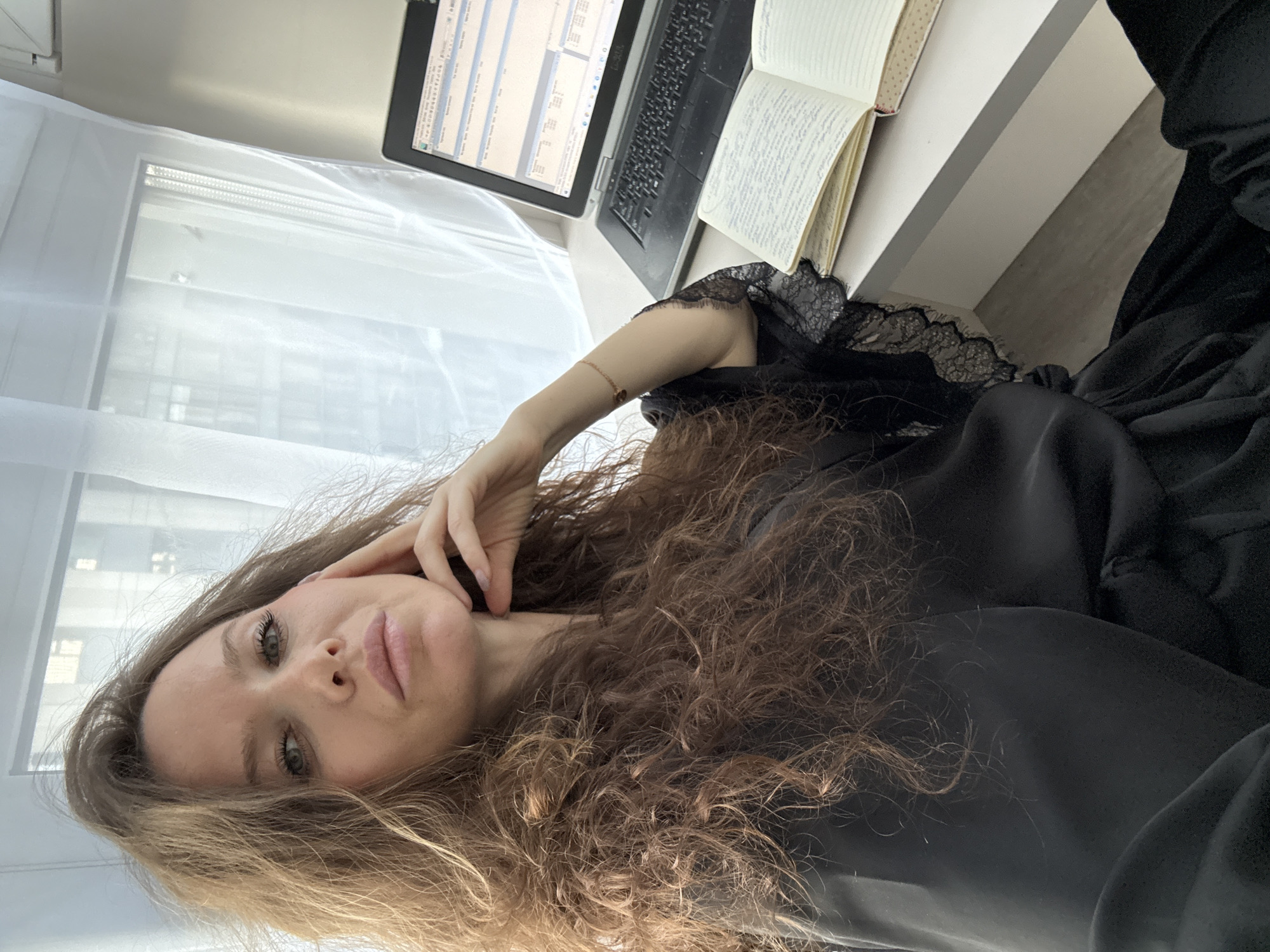 A young woman with long, curly, brown hair is pictured in a reclined or relaxed pose, looking directly at the camera. She has green eyes and a calm, possibly pensive, expression. Her left hand is gently resting near her jawline. She is wearing a black garment, which appears to be made of a soft, silky fabric, with intricate black lace trim visible on the shoulder/sleeve area. A thin, light-colored bracelet adorns her left wrist.
She is situated indoors, likely near a window, with sheer white curtains or blinds diffusing bright daylight into the space. A glimpse of what appears to be buildings, possibly in an urban or residential setting, can be seen through the window. Adjacent to her, on a white desk or surface, a black laptop is open, displaying a screen filled with what looks like a spreadsheet or data-intensive application. The "DELL" logo is visible on the laptop's bezel. Next to the laptop, an open notebook with handwritten text is present.
The scene suggests a moment of rest or a break from work or study, taking place during the daytime. No specific location or weather details are discernible beyond the indoor setting and natural light. The visible text includes "DELL" on the laptop and unreadable handwritten text in the notebook, along with unreadable digital text on the laptop screen.