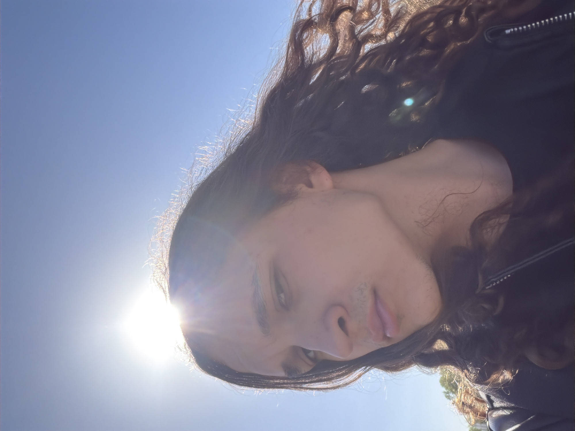 The image displays a close-up, rotated portrait of an individual with long, wavy dark hair and subtle facial hair, captured outdoors under a clear, bright blue sky. The subject's head is tilted significantly, appearing almost horizontal across the frame due to the image's orientation. They are wearing a dark jacket, possibly leather, with a zippered detail visible on the shoulder.
The scene is illuminated by intense sunlight, indicative of a clear, sunny day, likely midday or early afternoon. The sun is positioned behind and to the left of the person's head, creating a prominent lens flare and strong backlighting that outlines the individual's hair with a bright halo effect. This backlighting results in some natural shadowing on the face, though features like the eyes, nose, and lips are clearly visible. The person's expression is neutral, with a slight pout. While no specific architectural or geographical landmarks are visible, the location context provided indicates the image was taken in Plovdiv, Bulgaria. There is no visible text in the image.