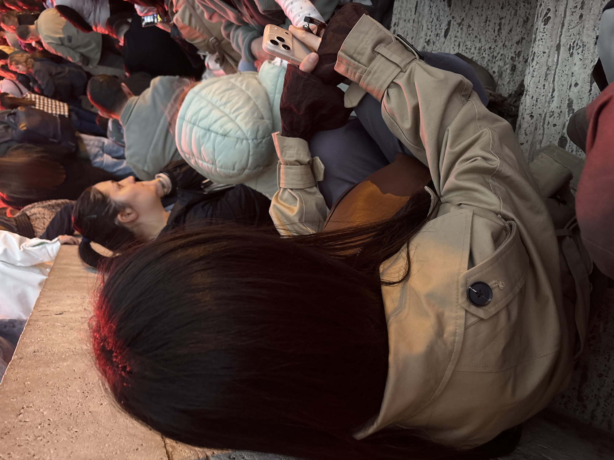The image captures a dense gathering of people in an outdoor urban setting, illuminated by artificial light, suggesting evening or nighttime. In the immediate foreground, a person with long, dark hair is seen from behind, wearing a beige trench coat with a visible dark button on a pocket. To their right and slightly behind, another individual's face is visible in profile, looking upwards, with dark hair tied back.
In the mid-ground, a hand wearing a dark glove or sleeve is holding a light-colored smartphone with multiple camera lenses, partially obscured by the sleeve of a beige jacket. Among the crowd, another person is distinguishable by a light mint green quilted jacket. The background is filled with numerous other individuals, forming a substantial crowd, dressed in various casual and warm attire.
The ground appears to be a light-colored concrete or stone surface where people are sitting or standing. The artificial lighting casts warm, reddish highlights on the subjects' hair and clothing, creating a distinct ambiance. The scene depicts a public event or social gathering in Yerevan, Armenia. No discernible text is visible within the image.