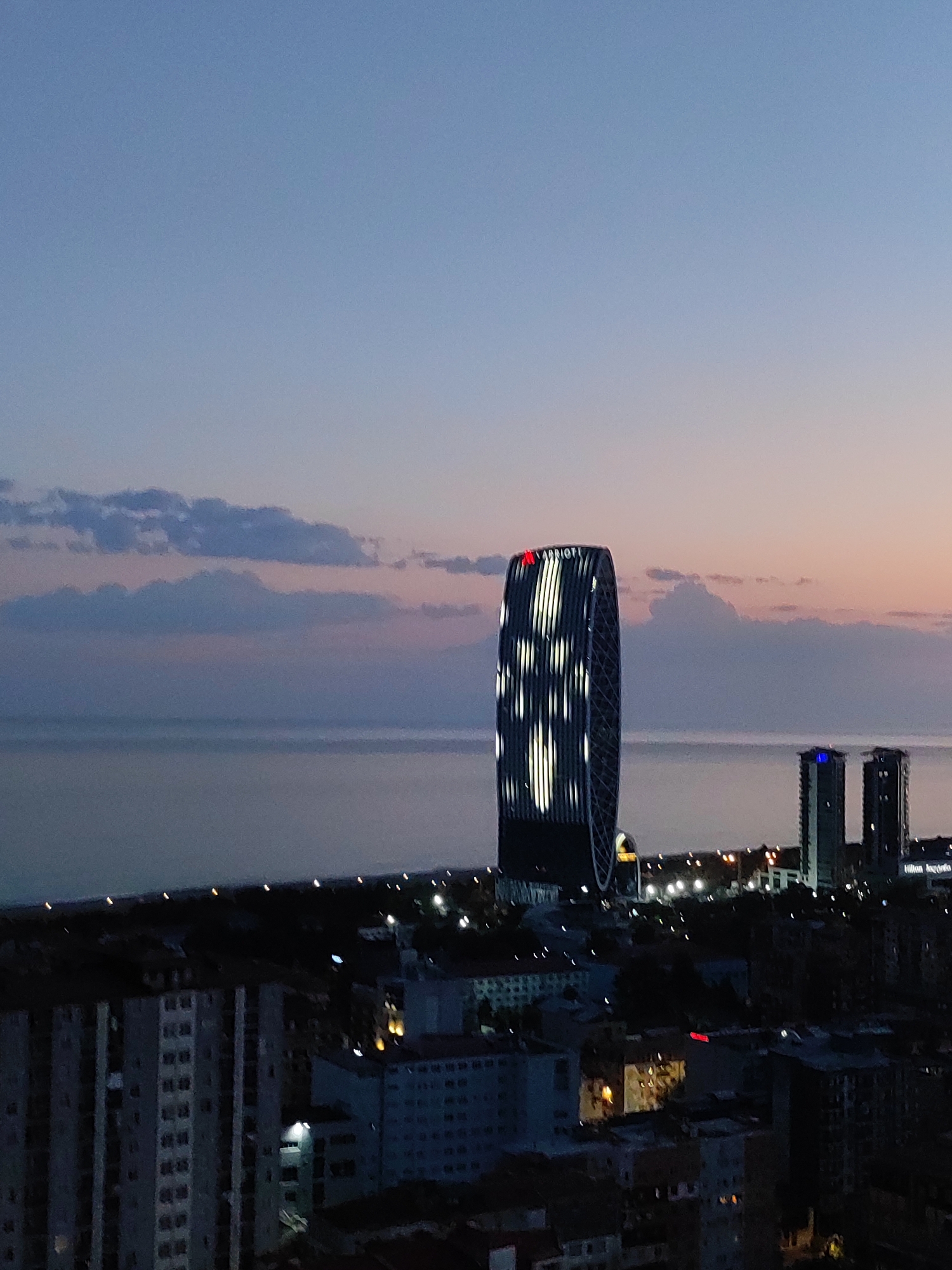 The image shows a cityscape at dusk, with the sky transitioning from a clear blue to shades of pink and orange near the horizon. A body of water, likely the Black Sea, is visible in the background. Several buildings are illuminated, with a distinctive, oval-shaped skyscraper prominently featuring the "MARRIOTT" logo at the top. This skyscraper has a patterned lighting design on its facade. To its right, two other tall buildings stand, one with a blue light on top. The foreground consists of a dense collection of lower-rise buildings, some of which are lit from within, suggesting it is a residential or commercial area. The overall impression is of a modern city at the edge of the sea as evening approaches.