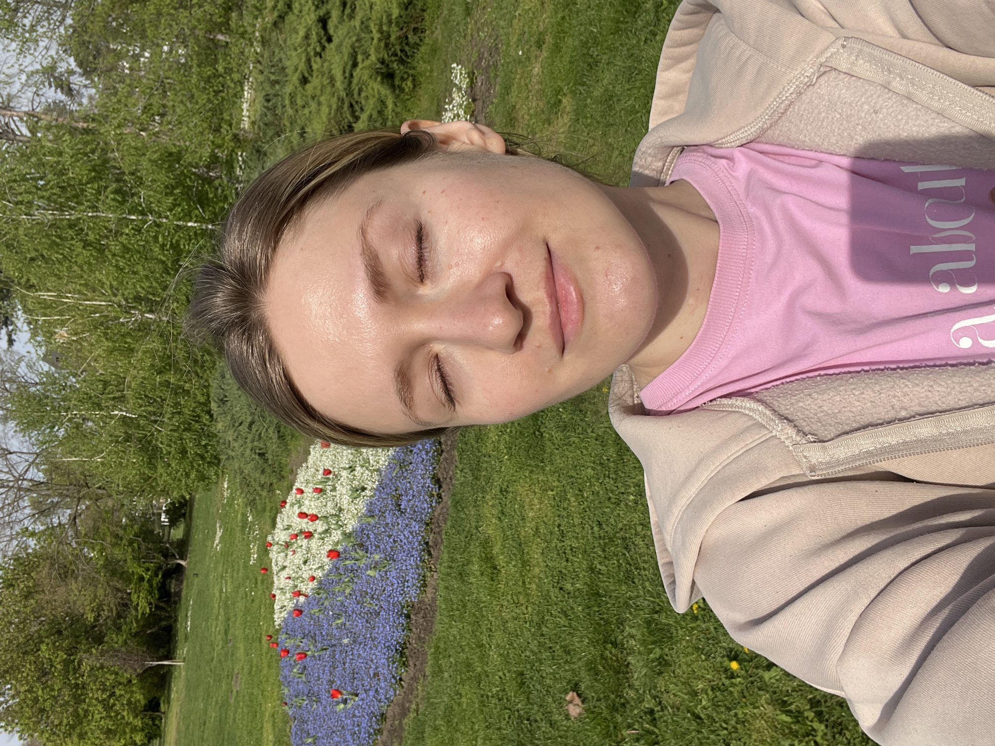 The image is a close-up selfie of a woman lying on green grass near a flowerbed. The woman has fair skin, light brown hair, and closed eyes. She is wearing a pink t-shirt with white text and a beige hoodie. The flowerbed has blue, white, and red flowers. The background features green trees and bushes. The location is a park in Novi Sad, Serbia.