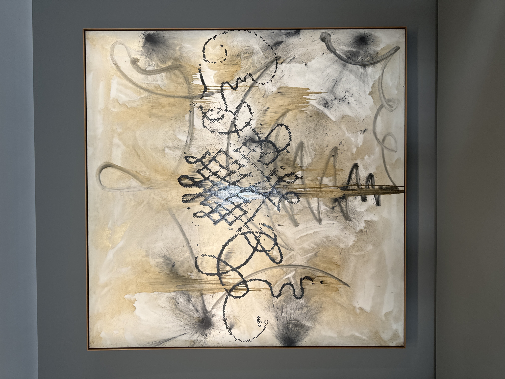 The media file showcases a square-format abstract painting mounted on a plain grey wall. The artwork features a dynamic composition rendered in a palette of muted tones, primarily consisting of various shades of beige, cream, white, black, and grey, with subtle shimmering accents of gold or bronze.
The background is characterized by layered washes and broad, expressive brushstrokes in lighter colors, creating a textured and somewhat ethereal base. Over this foundation, darker elements are applied with vigorous energy. A prominent central motif is formed by intricate, calligraphic lines composed of numerous black dots, resembling a complex knot, script, or abstract symbol. This central cluster is encircled by a network of sweeping, fluid lines and gestures in black and grey, varying from bold strokes to delicate, looping forms.
Several areas of the canvas display a distinct splattering or burst effect, particularly in black and grey pigments, contributing to the painting's energetic and spontaneous quality. The overall impression is one of rich texture, movement, and a harmonious balance between organic fluidity and structured yet abstract forms. The painting is enclosed by a thin, light-colored frame. No visible text is present within the artwork.