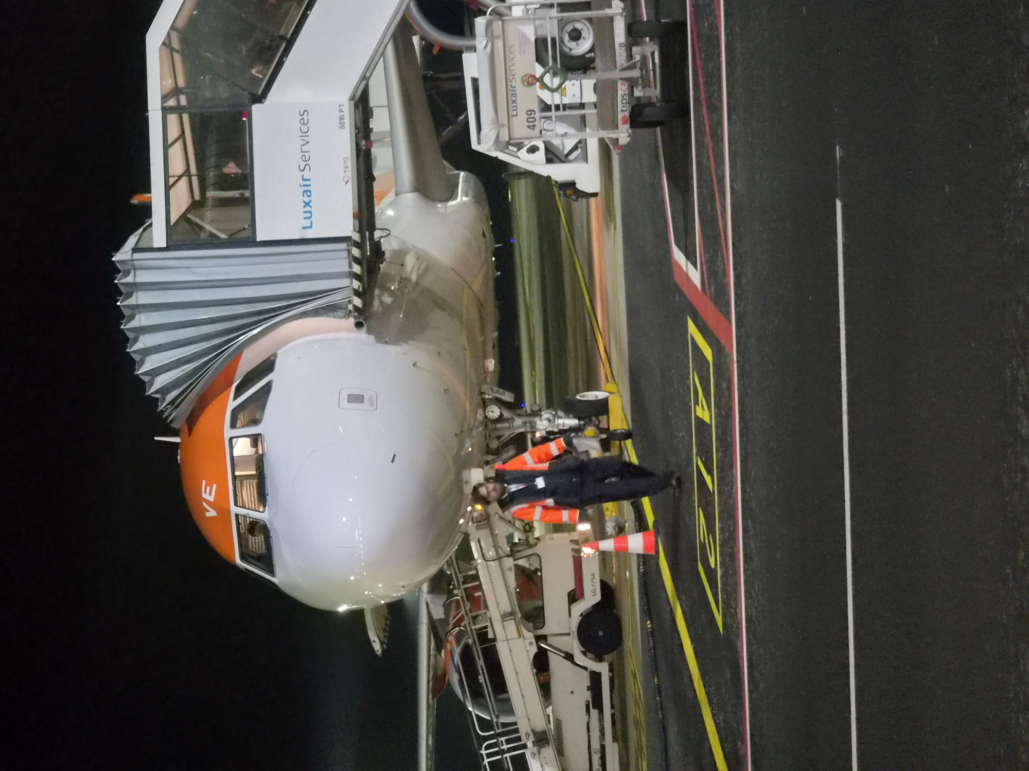 At an airport in Niederanven, Luxembourg, an EasyJet Airbus A320 aircraft is parked at a gate with a Luxair Services jet bridge connected. A ground crew member in a high-visibility jacket stands near the aircraft's nose and a ground support vehicle. The aircraft's registration "VE" is visible on the orange nose. The scene is illuminated under artificial lights at night. Markings on the asphalt indicate the aircraft's position, including yellow and white lines and the number "A13". A pushback tractor labeled "LuxairServices" is positioned near the aircraft's nose gear.