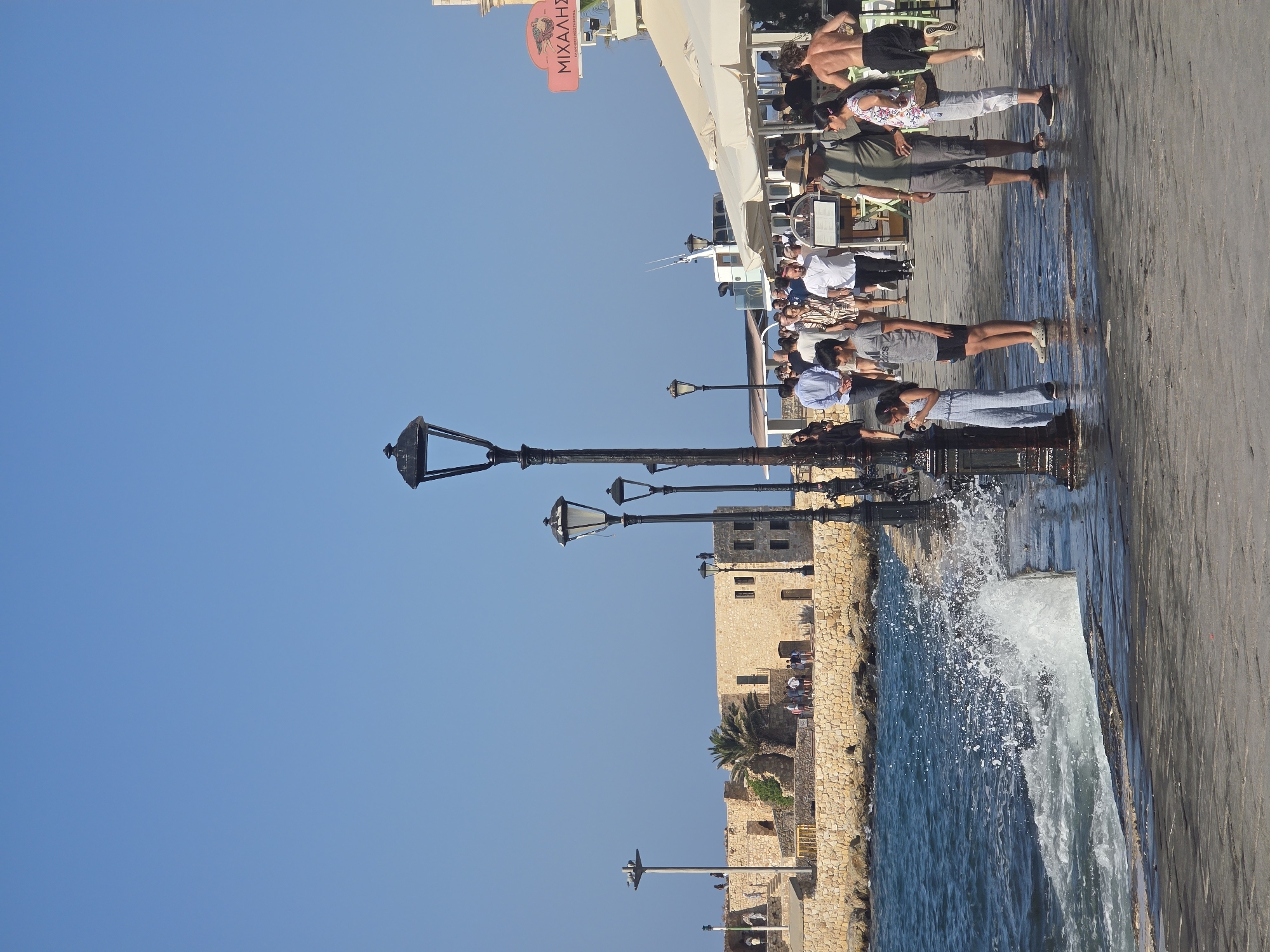 The image captures a lively waterfront scene in Chania, Greece, likely the Venetian harbor, on a bright, sunny day. A stone promenade lines the harbor, with people strolling along its edge. The water is a vibrant blue, with waves crashing against the walkway, creating a dynamic splash in the foreground.
Several people are visible, including families and tourists enjoying the seaside ambiance. A young girl in a light blue jumpsuit stands near a lamppost, looking down. Next to her, a boy in a gray t-shirt and black shorts is looking forward. Further down the promenade, a man in a green shirt and gray shorts stands with a woman wearing a floral-patterned shirt and white pants. To their right, a man with his back to the camera, wearing black shorts, is running.
In the background, a historic stone structure, possibly part of the Venetian fortifications, is visible with palm trees in front of it. Restaurants and cafes with outdoor seating and cream-colored awnings line the promenade, suggesting a bustling commercial area. A sign with Greek lettering, "ΜΙΧΑΛΗ'Σ," is mounted on a pole. Multiple ornate black lampposts are spaced along the walkway, adding to the historic character of the port. The clear blue sky suggests fair weather.