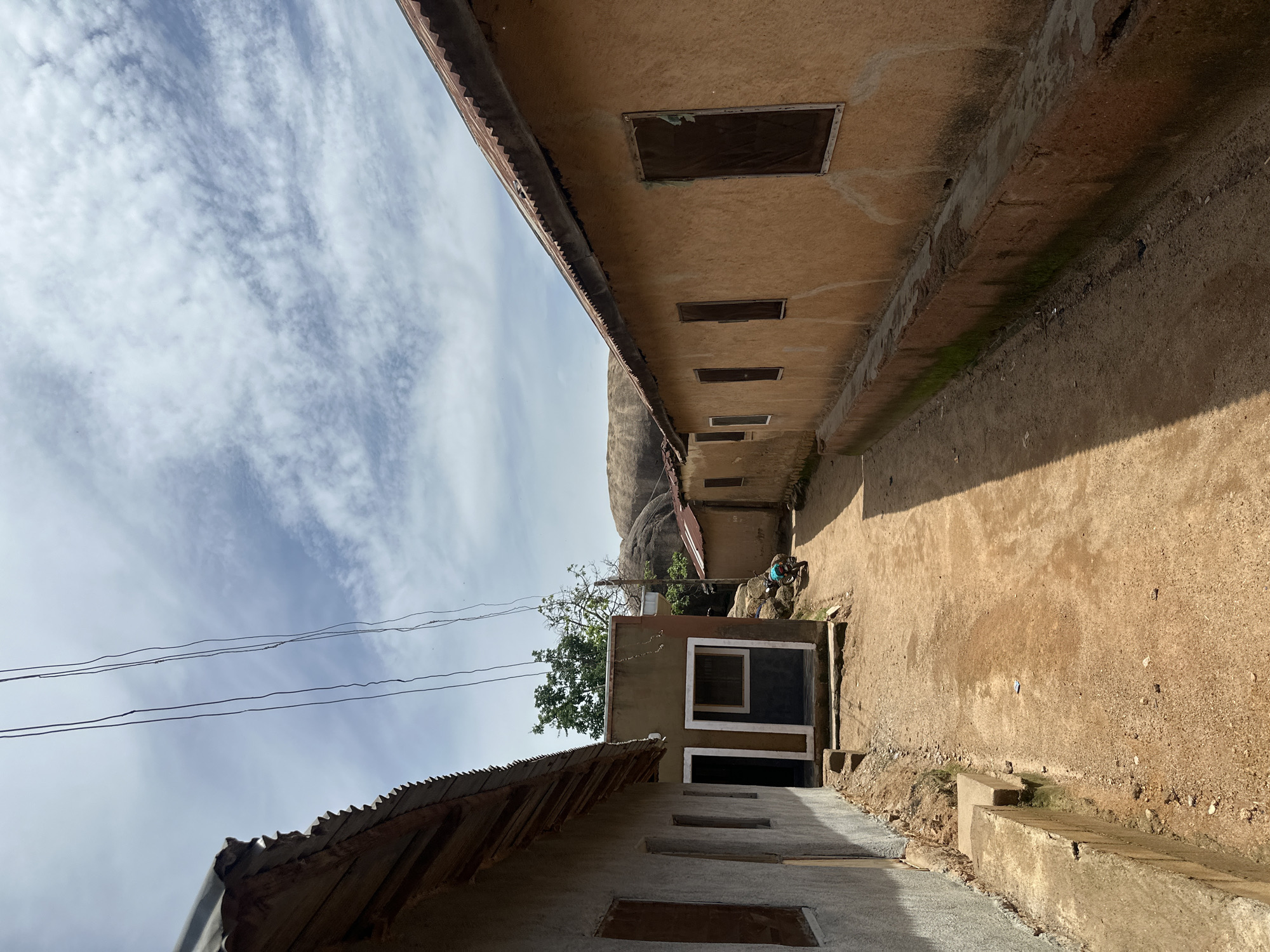 This is a ground-level, rotated image of a residential area in Jos, Nigeria. The scene features a narrow path between two buildings. The building on the right has a rough, textured, tan-colored facade with several small, rectangular windows. The building on the left, closer to the foreground, is partially constructed with a corrugated metal roof. Overhead, a bright blue sky is filled with wispy white clouds. Power lines are visible stretching across the sky.
In the distance, a large rock formation is visible, suggesting a hilly or mountainous terrain. A tree with green foliage grows near the rock formation. A small, single-story building with a darker colored section and a window is situated between the two main buildings. A person, possibly a child, wearing a blue shirt and dark pants, is kneeling or squatting on the ground near the rock formation, appearing to engage with something on the ground or rocks.
The sunlight casts strong shadows, indicating it is likely daytime, possibly late afternoon. The overall impression is of a simple, rural or semi-urban setting, with basic construction and a natural landscape. There is no visible text in the image.