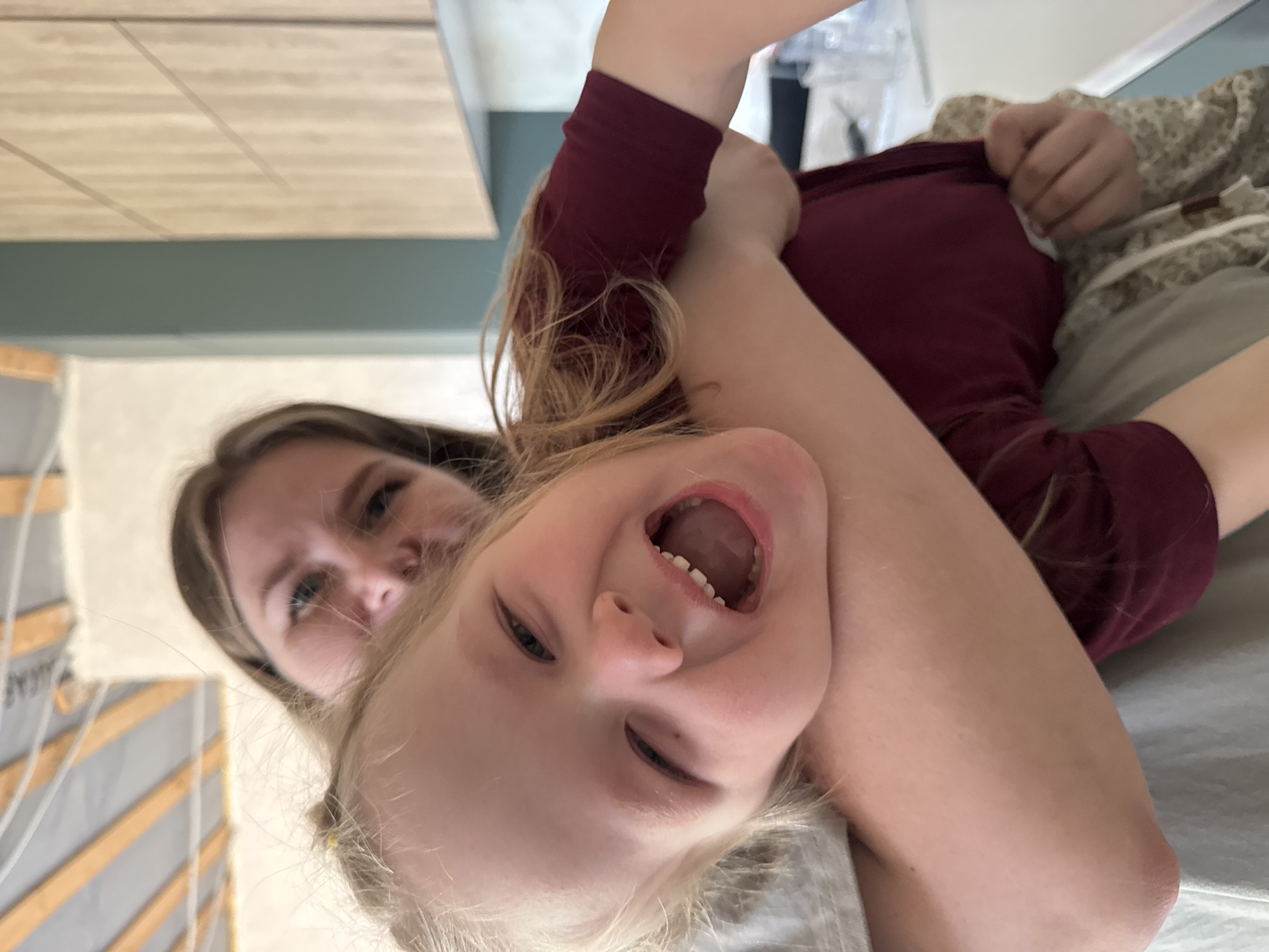 A woman and a young girl are in a close embrace. The girl is laughing with her mouth open, while the woman is smiling. They are indoors, the photo is taken at a tilted angle, capturing details of the room: wood texture, the color gray. The location is Gornyi, Dvurechenskoe S/P, Russia.