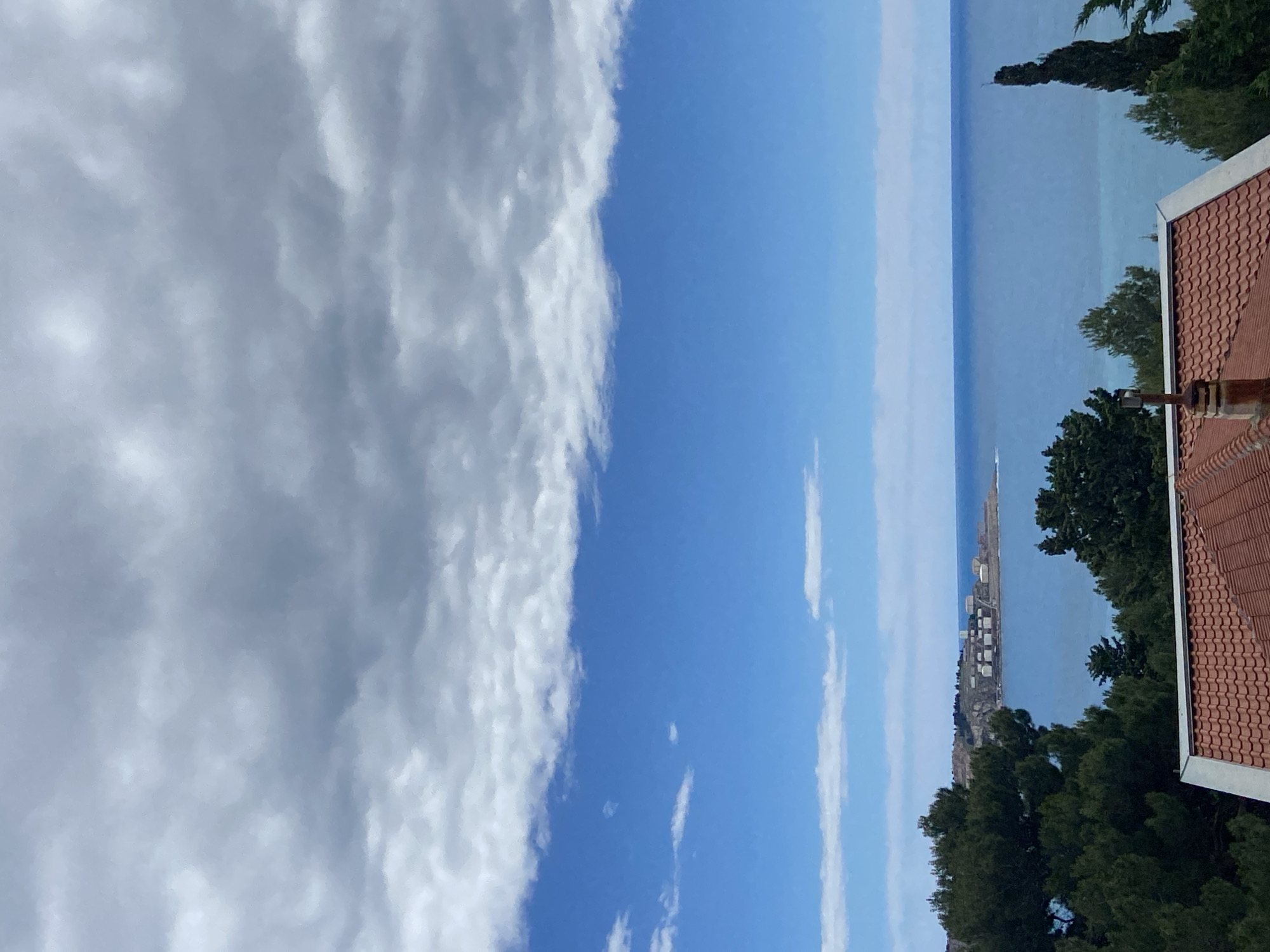 The image shows a view from an elevated position in Šušanj, Montenegro, overlooking the Adriatic Sea. A dense, cumulonimbus cloud formation dominates the left side of the frame, contrasting with the clear blue sky and sea towards the right. A narrow strip of land, potentially a peninsula or breakwater, is visible in the distance. In the foreground, the roof of a terracotta-tiled building is partially visible, framed by green trees, suggesting a residential area. The scene is still and quiet.