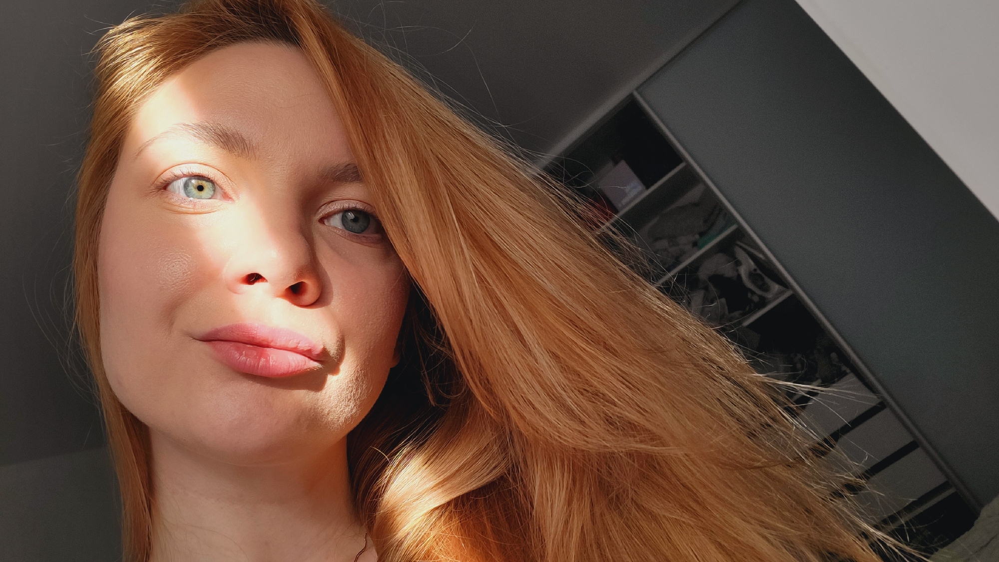 A fair-skinned woman with strawberry blonde hair is the subject of this close-up photograph. Light casts shadows across the left side of her face, illuminating one eye as well as her nose and the side of her lips. The background includes what appears to be a modern wardrobe or closet with dark grey doors, featuring shelves visible within the open compartments. While the image offers no clear indicators of the exact location, the prompt suggests that the photo was taken in the city of Chaikovsky, Russia.