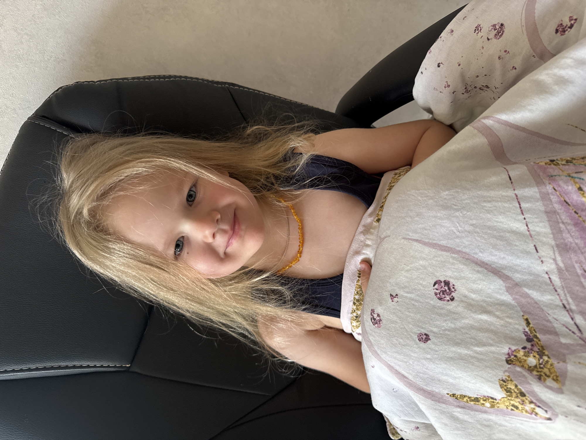 A young girl with blonde hair and blue eyes is depicted in a diagonal, close-up shot, lying on her side and looking directly at the camera with a soft, gentle smile. Her head rests on a black leather chair, detailed with white stitching. She wears a dark blue top and an amber beaded necklace. A light-colored blanket or duvet, featuring a pattern of purple and gold accents, covers a portion of her body. The background consists of a plain, light-toned wall, suggesting an indoor setting. The scene conveys a moment of calm and relaxation. The location context provided is Ферма, Russia, though no specific external cues for this location are visible in the indoor shot. No visible text is present in the image.