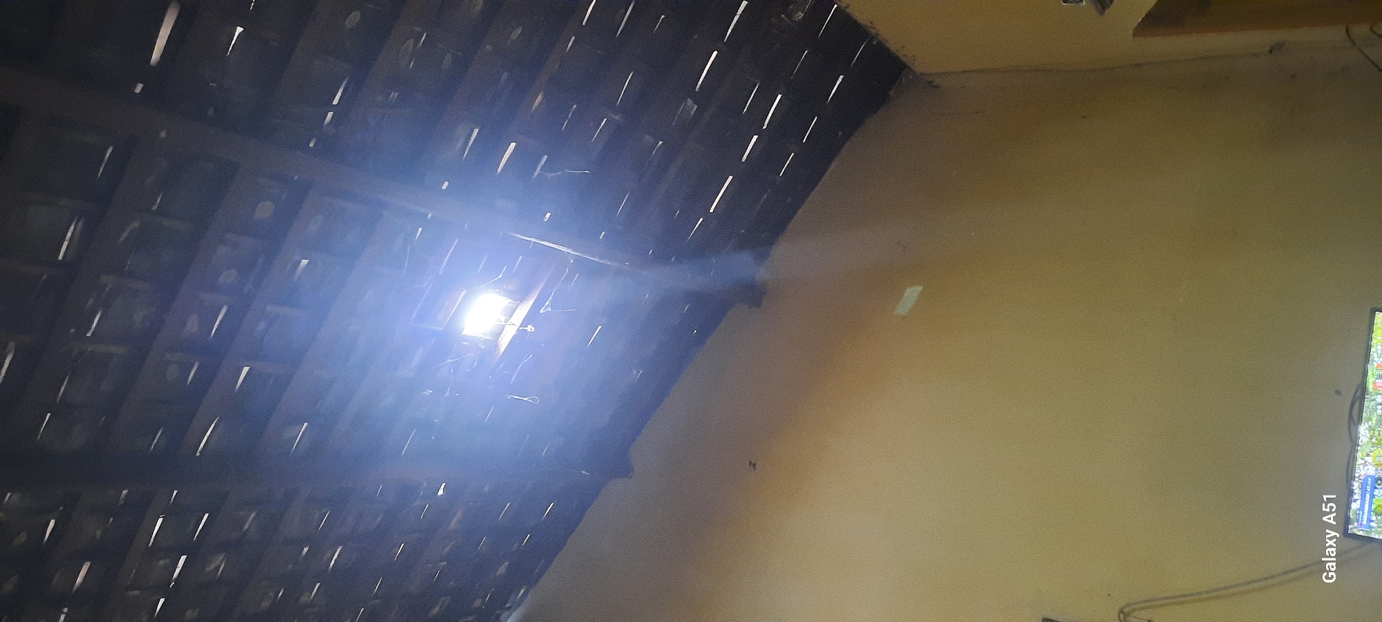 The image depicts the interior of a room looking upwards towards a partially open roof or ceiling. Light beams penetrate through gaps in the dark, tiled roofing structure, illuminating dust particles in the air. The walls of the room are a light yellow color. In the bottom left corner, a Samsung Galaxy A51 watermark is visible, suggesting the photograph was taken with this device. A small portion of a television screen is also visible at the very bottom of the image, displaying a colorful image. The scene is domestic and appears to be an indoor space in Ploso, Indonesia.