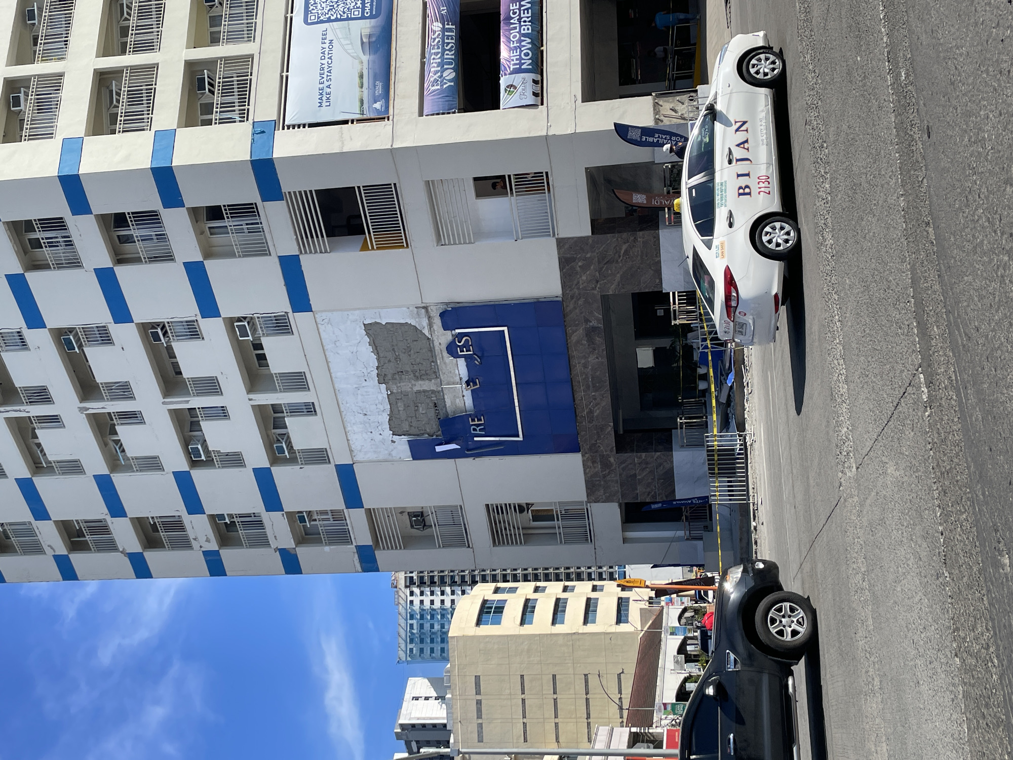 The image captures a street scene in Davao City, Philippines, in front of a multi-story building. The building features a facade with alternating white and blue square patterns, and several balconies with white railings are visible. A portion of the building's exterior appears damaged, with exposed concrete and rebar. Below the damaged section, a blue sign with white letters that partially spells "RE ES" is mounted. Two large banners are affixed to the building: one reads "MAKE EVERY DAY FEEL LIKE A STAYCATION" and another, partially obscured, has text related to "EXPRESS YOURSELF" and "THE FOLIAGE".
In the foreground, a white taxi with the branding "BIJAN" and the number "2130" is driving on a paved road. To the left, a black SUV is also on the road. Yellow caution tape is visible across the sidewalk and entrance to the building, suggesting ongoing construction or a restricted area. Several people are present near the building entrance and in the background, appearing to be either workers or passersby. The sky is bright blue with scattered clouds. Other buildings, including taller structures with glass facades, are visible in the background, indicating an urban environment.