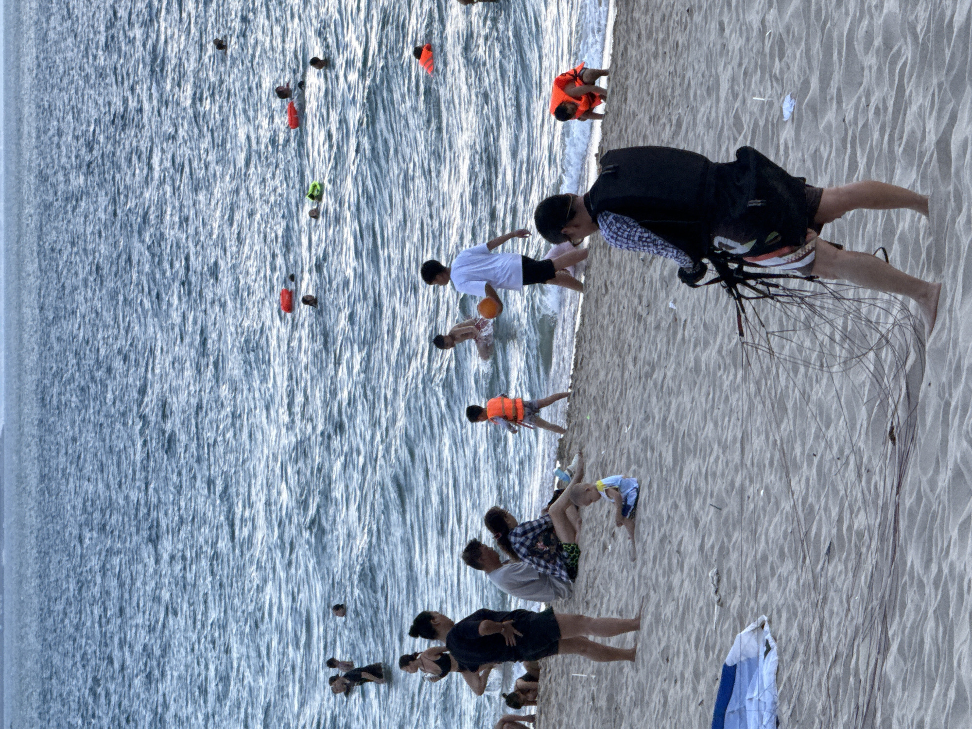 The image shows a crowded beach in Nha Trang, Vietnam. Many people are in the water, some swimming and others wearing life vests. On the sand, a few families are relaxing, and children are playing. In the foreground, a person is standing with what appears to be paragliding equipment. The weather seems to be overcast, and the sea is calm with moderate waves.