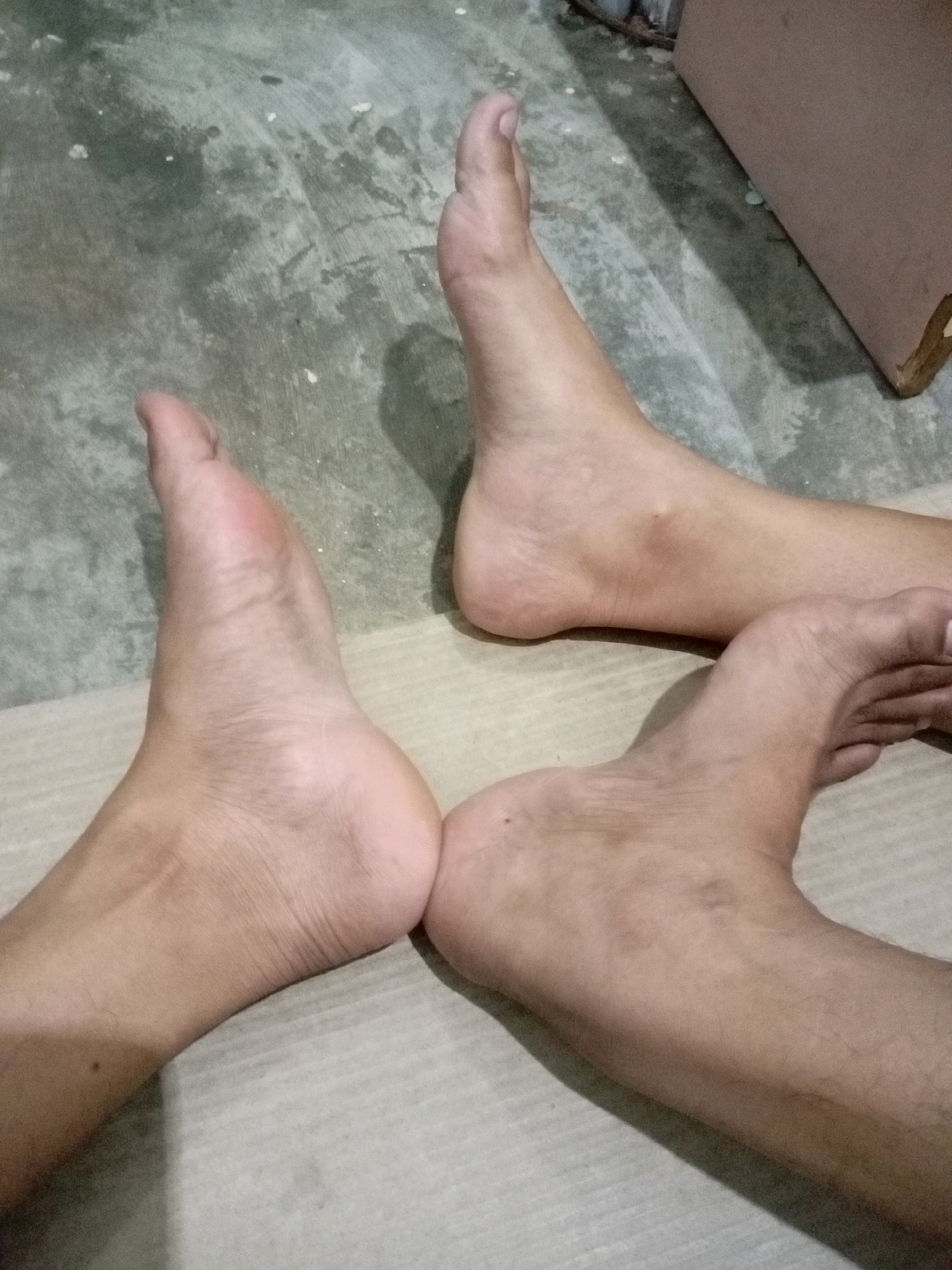 The image depicts a close-up, overhead view of three human feet positioned on a concrete floor with a textured surface. The feet are bare and show varying skin tones. One foot, on the left side of the frame, is positioned with its heel facing upwards and toes pointing towards the center. Another foot is placed to its right, with the heel closer to the viewer and the toes angled slightly upwards. A third foot is positioned above and to the right of the other two, with its heel further back and toes pointing downwards. The lighting casts shadows on the floor, creating a sense of depth. The background is out of focus but appears to be an indoor environment with a wooden object visible on the right side. The scene is devoid of any specific identifiable landmarks or clues that would indicate its location in Batam, Indonesia.