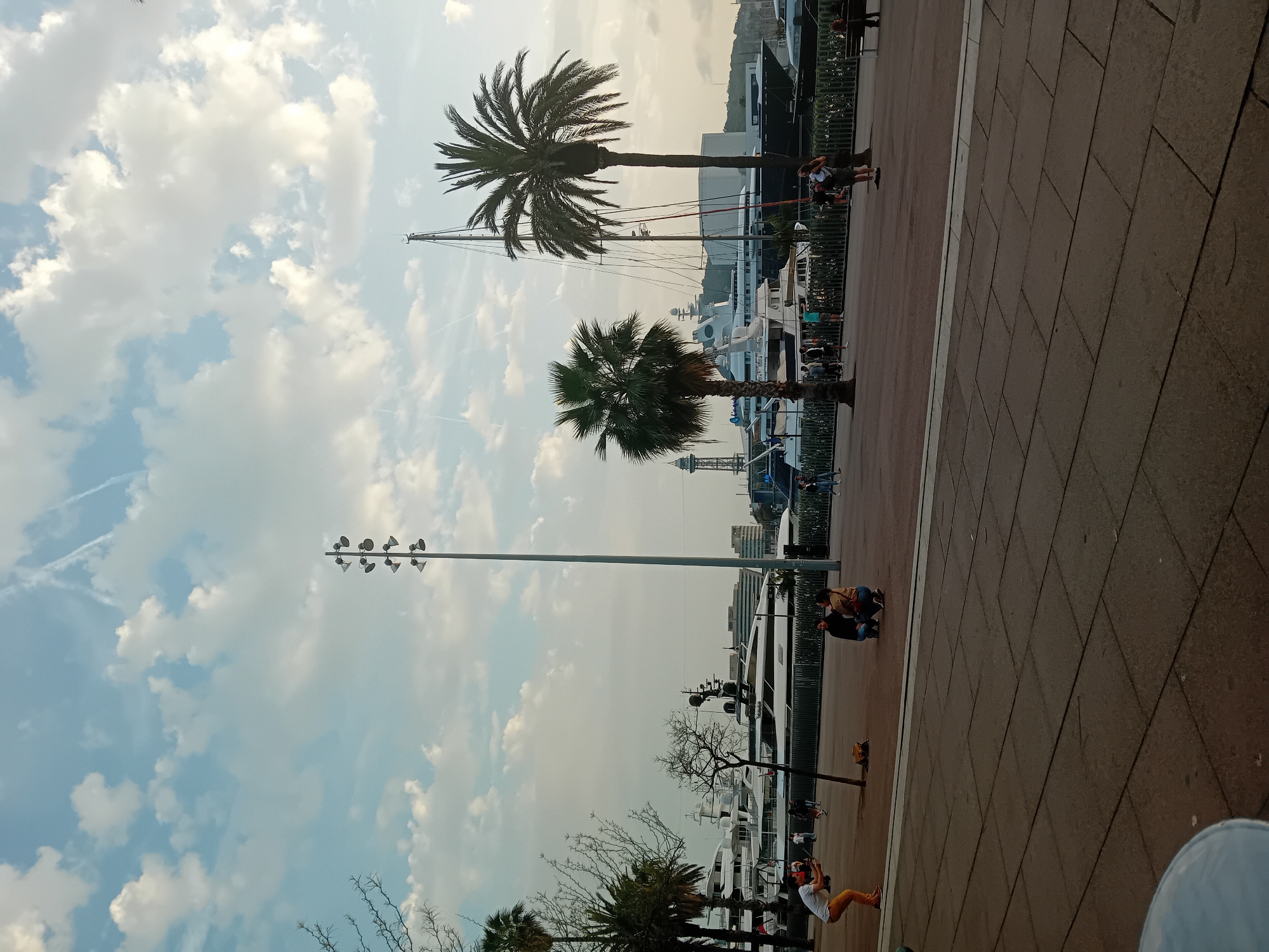 The image captures a vibrant waterfront scene, likely a promenade or marina in Barcelona, Spain. The scene is dominated by a wide, reddish-brown paved walkway in the foreground and mid-ground, bordered by a darker fence. Beyond the fence, numerous large, white yachts and boats are docked along the water.
Several people are present, engaging in various leisurely activities. In the lower left, a person wearing a white shirt and yellow/orange pants is crouched down, possibly taking a photo or looking at something. Further right, two individuals are also crouched together. Other smaller figures can be seen walking or standing near the boats in the background.
Two prominent palm trees stand in the mid-ground, with another smaller palm tree visible. A tall, slender lamppost with multiple light fixtures extends upwards from the left side of the frame. The sky is bright with scattered white cumulus clouds, and faint airplane contrails are visible. The lighting suggests late afternoon or early evening, with a warm, soft glow in the sky transitioning from a yellowish hue near the horizon to a lighter blue overhead. The overall atmosphere appears relaxed and pleasant. No discernible text is visible in the image.