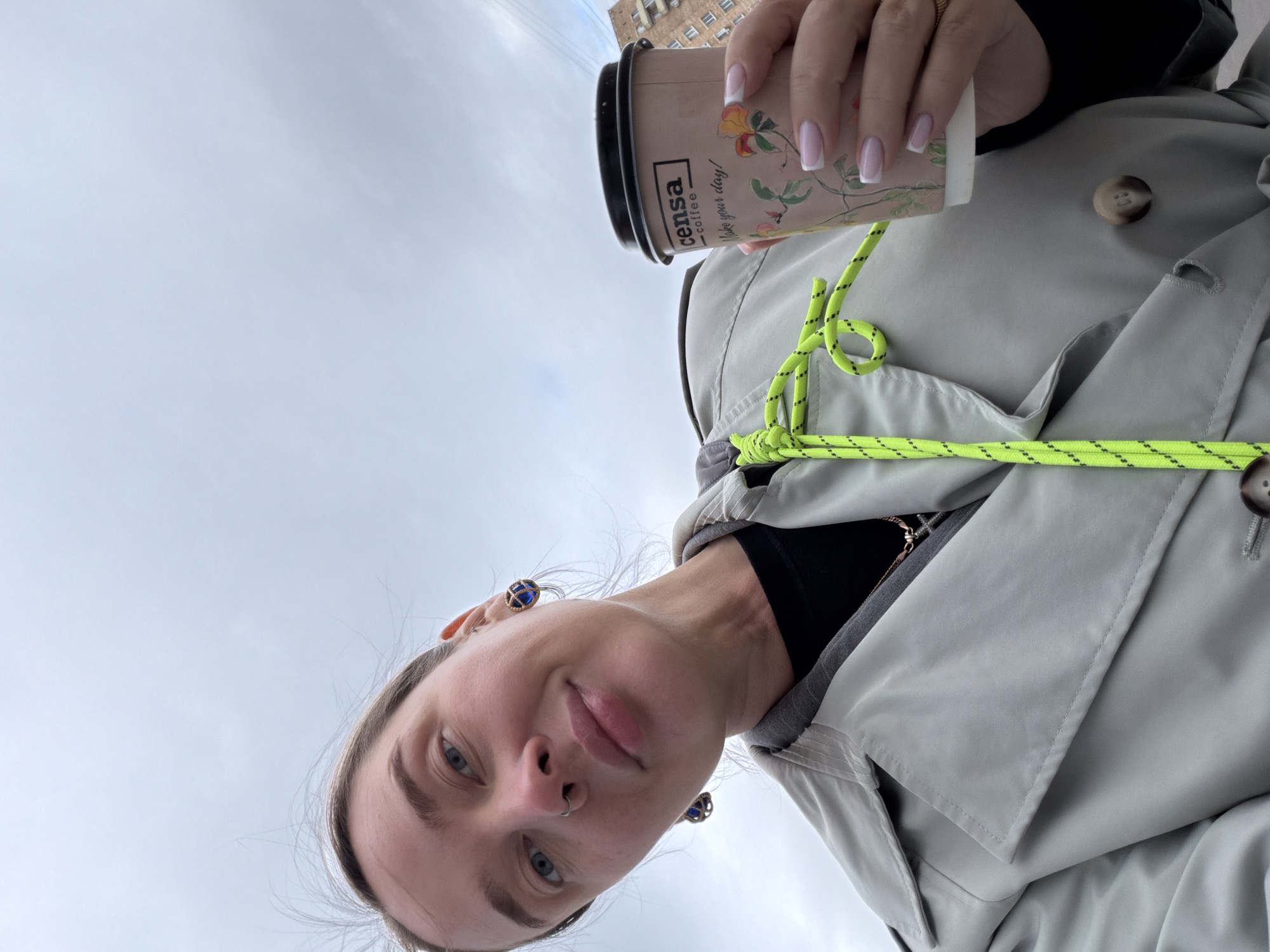 A photograph taken in Moscow, Russia, shows a person holding a coffee cup. The cup is labeled with "censa coffee" and the phrase "make your day!". The person is wearing a light gray coat with visible buttons and a neon green rope detail. The person has light skin, blue eyes, and a nose ring. Buildings are visible in the background. The sky is overcast.