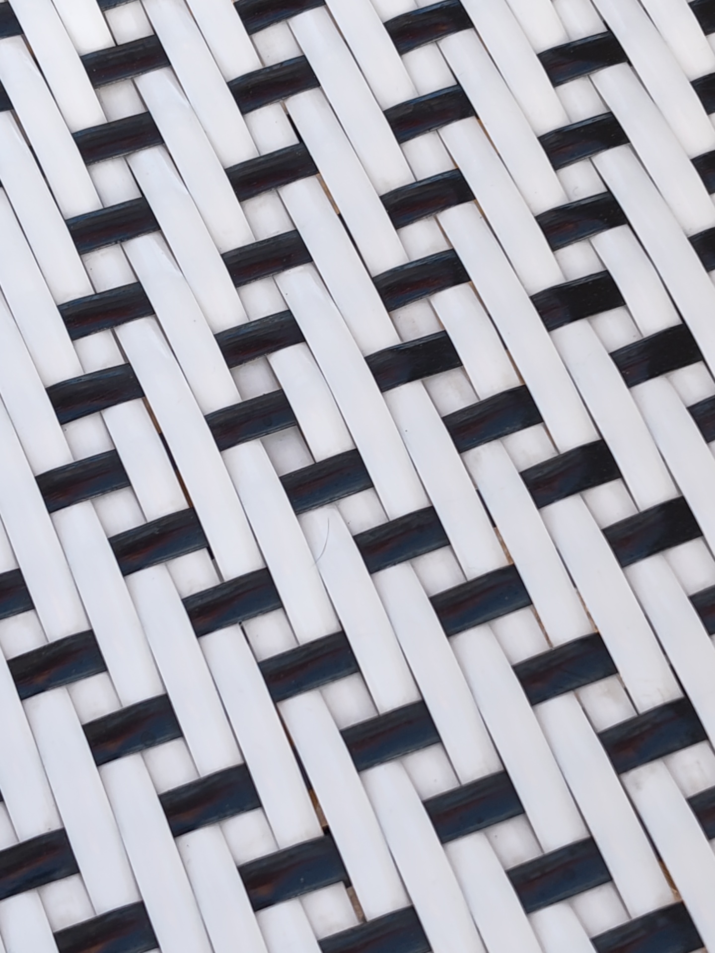 The image displays a close-up, top-down view of a woven surface, located in Dénia, Spain. The surface features an interlocking pattern of alternating white and dark-colored strips, likely black or very dark blue, arranged diagonally across the frame. Each strip is flat with slightly rounded edges, woven in an over-under configuration to form a dense grid. The material appears synthetic, exhibiting a slight sheen. Minor surface wear is visible, including faint scuffs and slight discoloration on some white strips. A light brown or reddish-brown hue is discernible through the small gaps in the weave. No individuals, actions, or specific environmental context beyond the woven object itself are present.