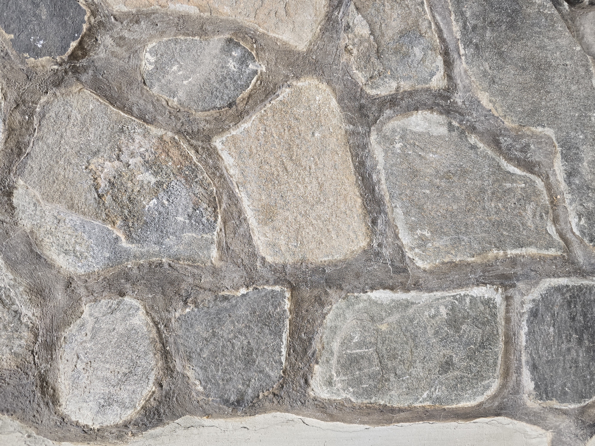 The image displays a close-up, eye-level view of a stone wall or paved surface. The primary subject is a rustic arrangement of natural, irregularly shaped stones set in mortar.
The stones vary significantly in size and shape, lacking uniform cuts or patterns. Their coloration is predominantly gray, encompassing a spectrum from light silvery gray to dark charcoal. Interspersed among the gray stones are several that exhibit warmer tones, including light beige, sandy brown, and hints of reddish-brown, suggesting different mineral compositions or weathering. The surfaces of the stones show varied textures, some appearing relatively smooth while others are distinctly rough and fractured.
Dark gray to brownish-gray mortar fills the wide, winding gaps between the individual stones. This mortar has a coarse, uneven texture and appears somewhat recessed, which accentuates the irregular outlines and three-dimensional quality of the stones.
At the bottom edge of the image, a horizontal strip of a distinctly lighter, smoother material is visible, stretching across the entire width. This material is light gray, possibly concrete or stucco, and forms a base beneath the stone work. Fine, hairline cracks are discernible on the surface of this lighter strip.
The scene depicts a static architectural detail. There are no people, activities, or recognizable objects beyond the construction materials. The lighting is even, highlighting the textural details of both the stones and the mortar. No text is visible in the image.