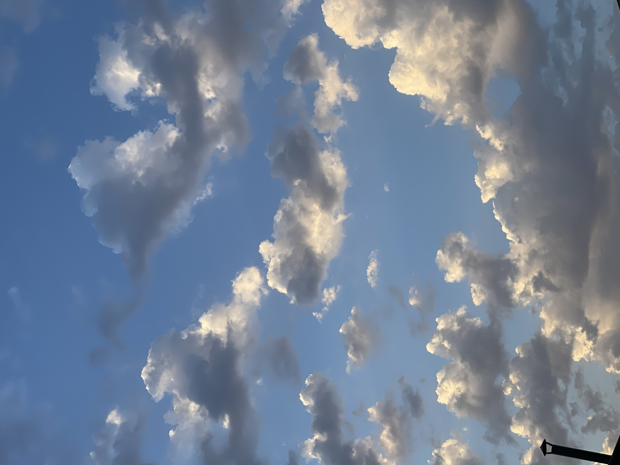 The image shows a blue sky with a scattering of cumulus clouds. The clouds are a mix of white and gray, with some illuminated by the sun and others in shadow. In the bottom left corner, a portion of a dark-colored, slender, vertical structure with a conical top is visible, possibly part of a chimney or a lamppost, against the sky. The scene is an outdoor view, likely from a high vantage point or an open area. There are no people or animals visible in the image. The time of day appears to be late afternoon or early evening, given the lighting conditions on the clouds. The photograph is taken from a vertical perspective, looking upwards at the sky. The location in Tashkent, Uzbekistan cannot be determined from the visual information provided in the image.