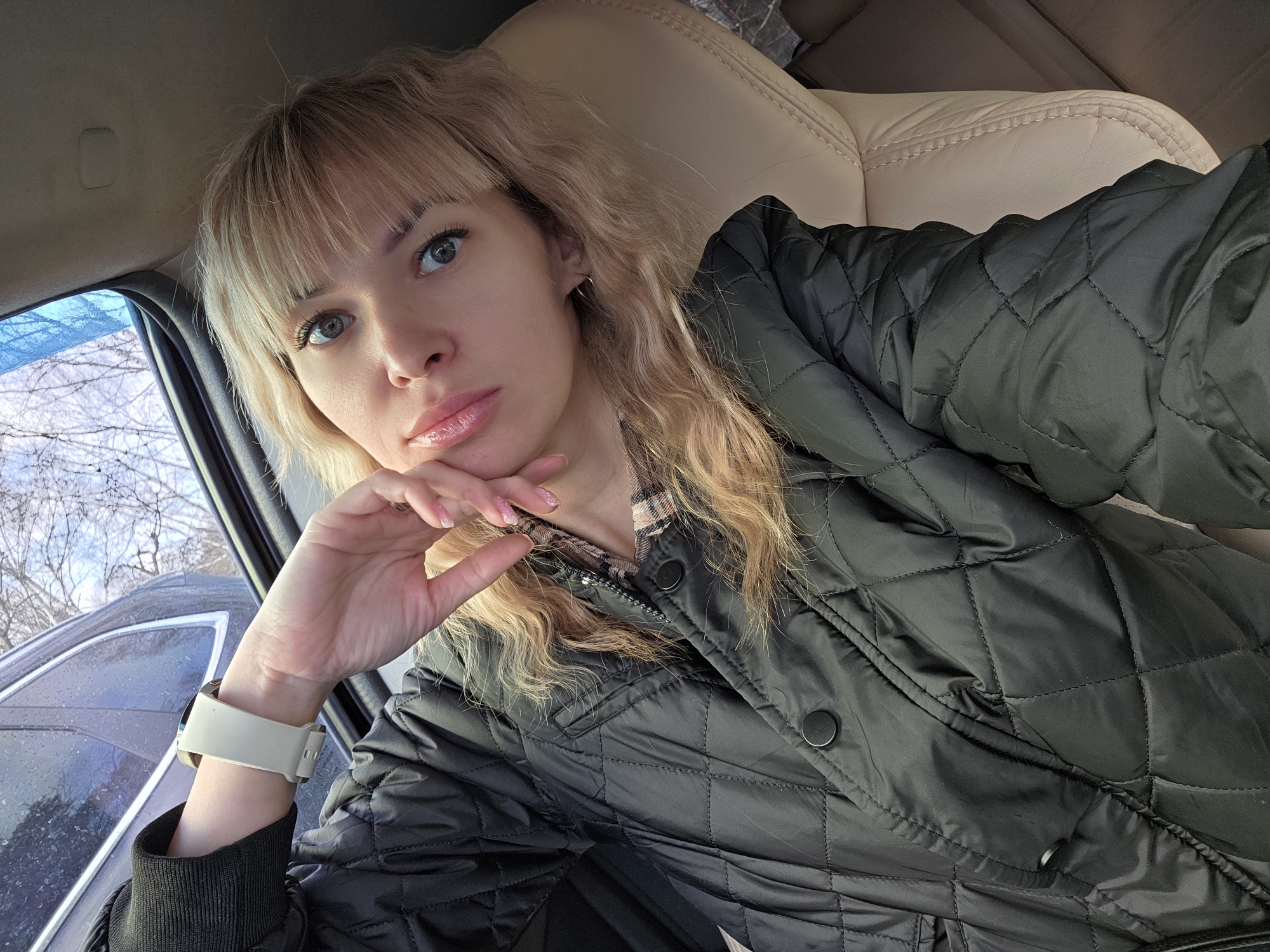 A woman with blonde, wavy hair and bangs takes a selfie from inside a car. She has light-colored eyes and full, glossy lips, maintaining a direct and slightly pensive gaze towards the camera. She is wearing a dark, possibly black or dark green, quilted jacket, under which a plaid shirt with pink and brown tones is visible near her collar. On her left wrist, she sports a white smartwatch. Her left hand rests gently under her chin, showcasing light pink, glittered nail polish.
The setting appears to be the interior of a car, with a light beige leather car seat visible behind her, suggesting a comfortable, personal space. Through the car window on the left, bare tree branches are visible against a bright, possibly overcast sky, indicating a cool season such as winter or early spring in Chaikovsky, Russia. The natural light suggests it is daytime. No discernible text is visible within the image.