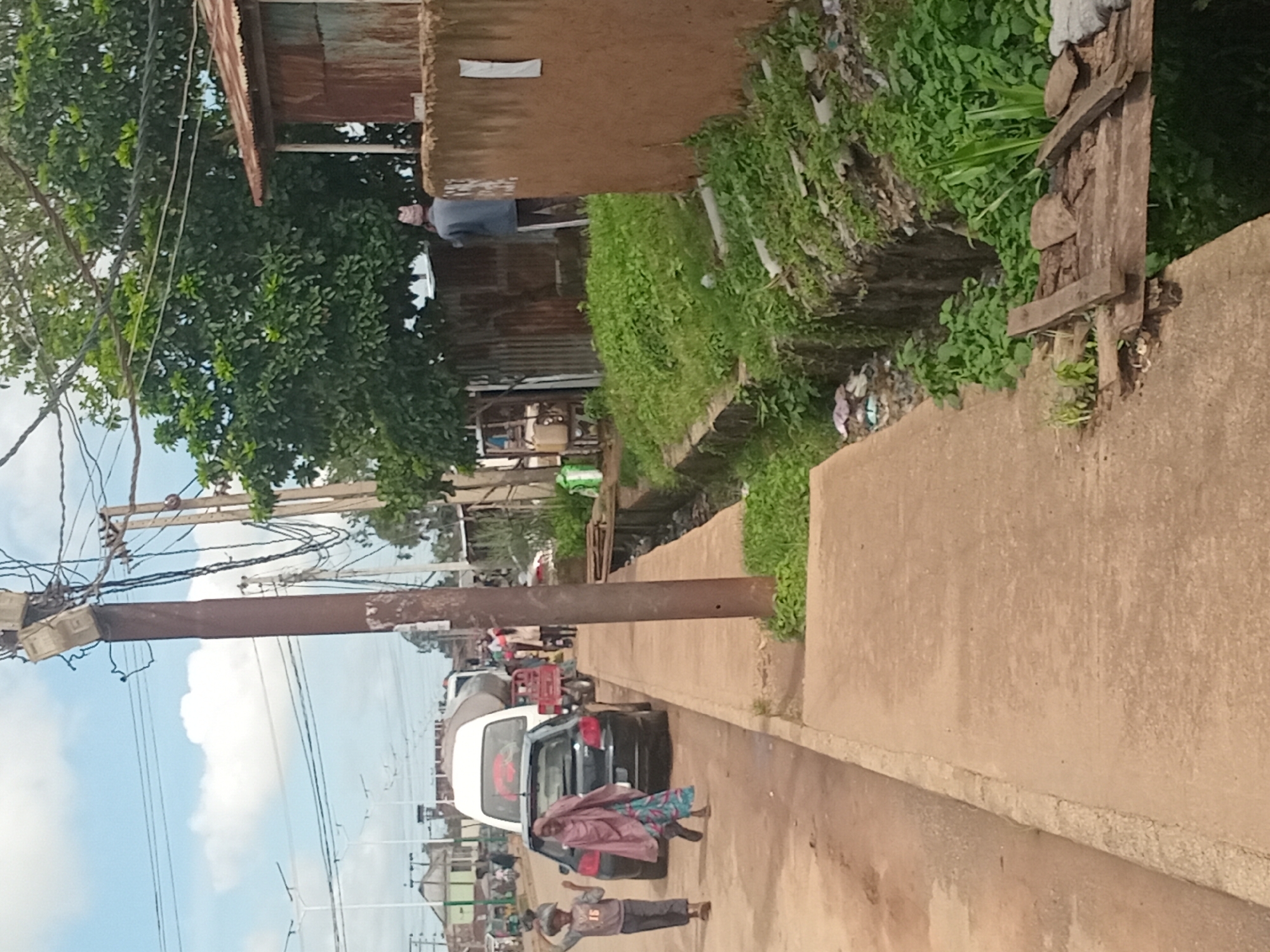 A street scene in Bukuru, Nigeria features a dirt road alongside a concrete sidewalk. On the road, several vehicles are present, including a black car, a white truck with a tank, and a small red motorcycle. Pedestrians are also visible, with two children and an adult woman walking on the sidewalk near the car. Further down the road, another person is walking away from the camera.
To the right of the sidewalk, a drainage ditch runs along the side of the road. This ditch is filled with green vegetation and debris, suggesting it is not well-maintained. A makeshift wooden bridge crosses the ditch in one section. Adjacent to the ditch is a brown, mud-brick wall with a rusted corrugated metal structure behind it. A tree with green foliage grows behind the metal structure.
Overhead, power lines crisscross the sky, supported by wooden and metal poles. One pole in the foreground has several electrical meters attached to it. The sky is partly cloudy. The overall impression is that of a busy, developing urban environment with some signs of disrepair.