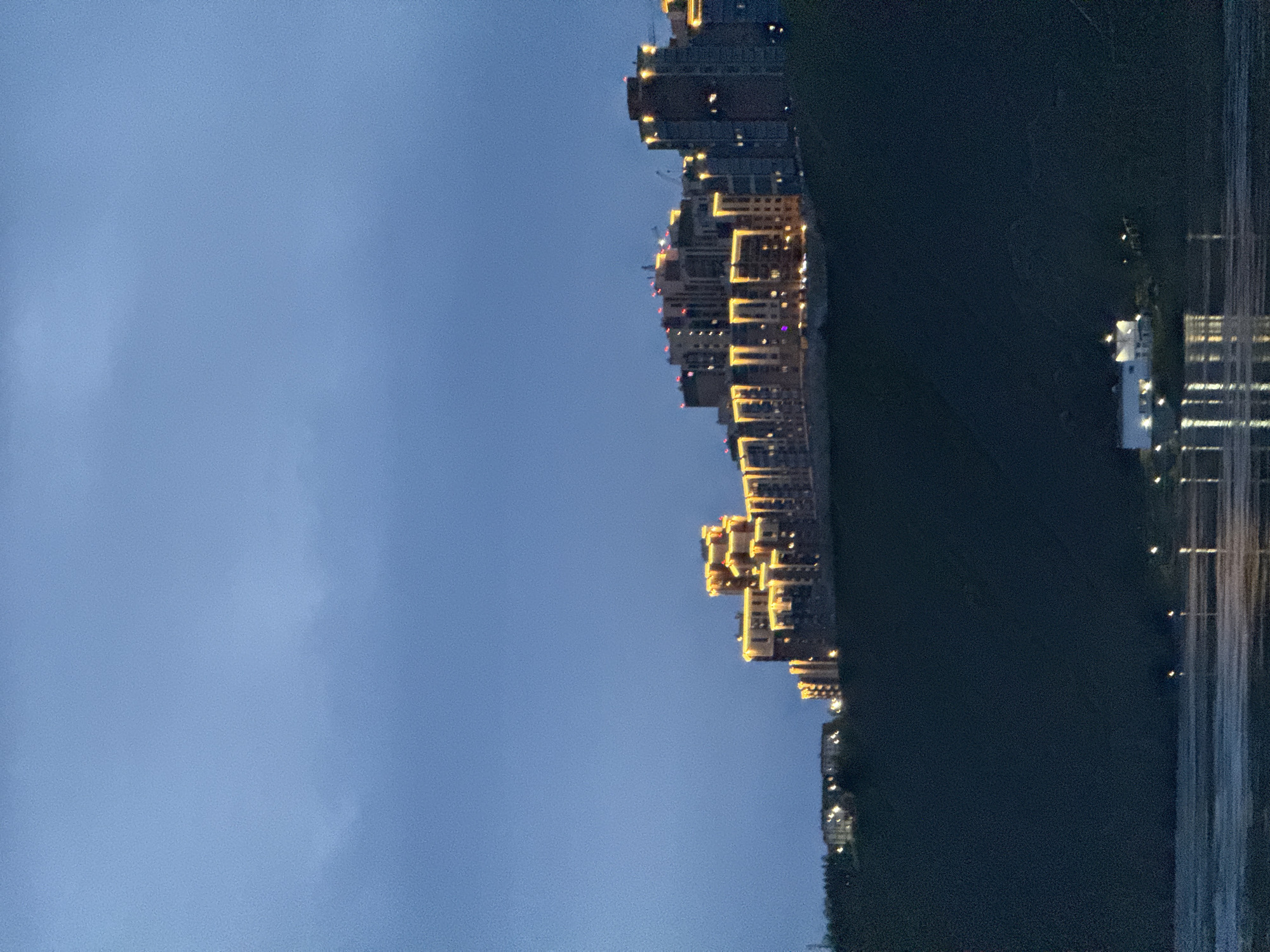 A long exposure photograph captures an urban landscape at dusk. Tall, modern buildings with illuminated windows stand along the edge of what appears to be a body of water, their lights reflecting in the water. The sky transitions from light blue to a darker blue near the top of the frame. Trees line the right side of the buildings. The photo is oriented such that the sky appears to be on the left side and the buildings run vertically along the upper part of the frame. The location may be Krasnoyarsk, Russia.