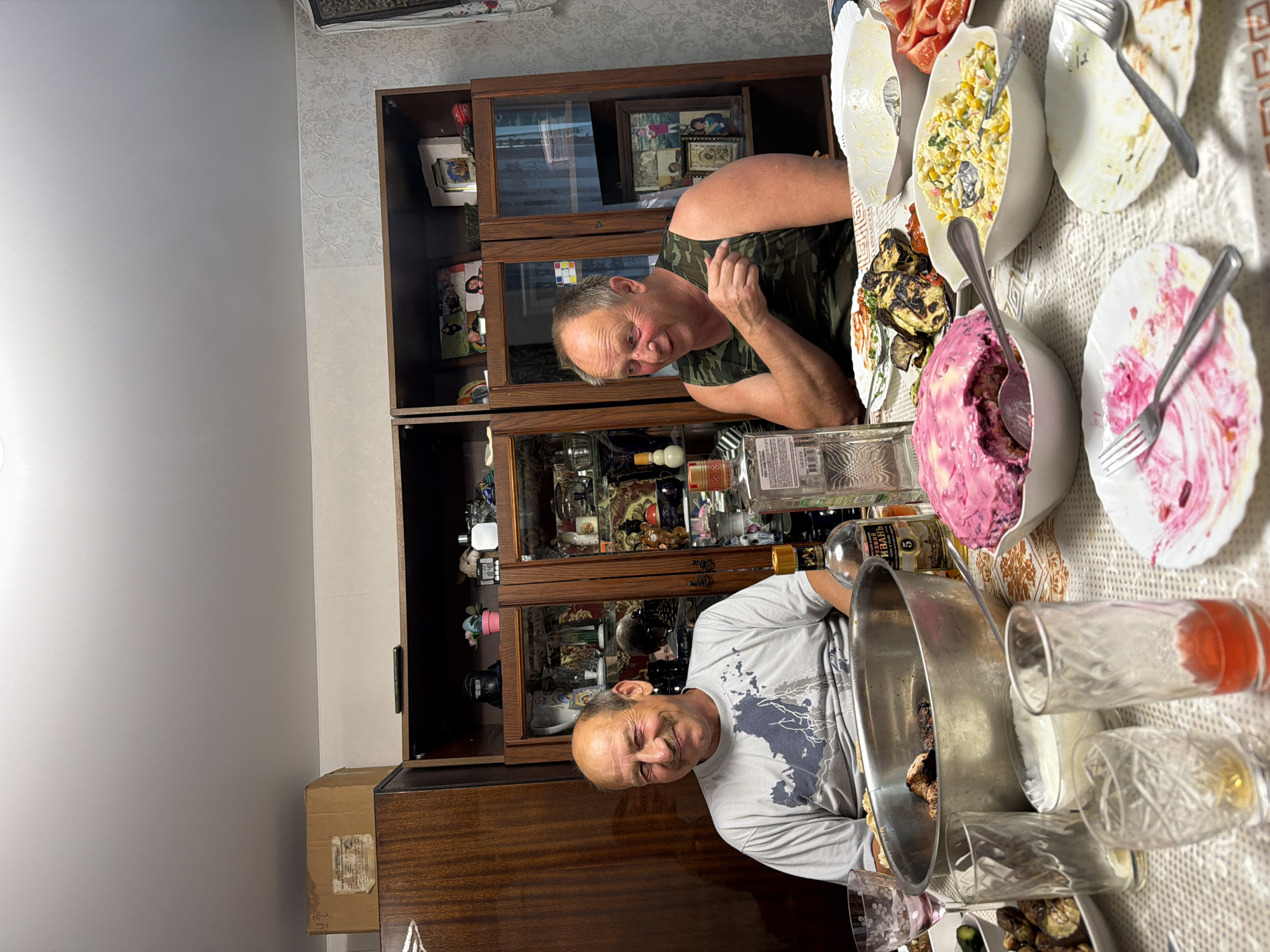 Two men are seated at a dining table laden with food, appearing to be in a residential dining room, possibly in the city of Бронница, Russia. The man on the left, wearing a grey t-shirt with a blue graphic, is smiling and looking towards the camera. The man on the right, in a camouflage tank top, is gesturing with his right hand and looking to his left. The table is covered with a patterned tablecloth and features several dishes of food, including a large bowl of meat, a pink layered salad, a corn and vegetable salad, grilled vegetables, and sliced tomatoes. Several empty and partially filled glasses are also visible, along with bottles of alcohol. In the background, a wooden cabinet with glass doors displays various items, and framed photographs are visible on top of the cabinet. The room appears to be lit, with a ceiling light possibly contributing to the illumination.