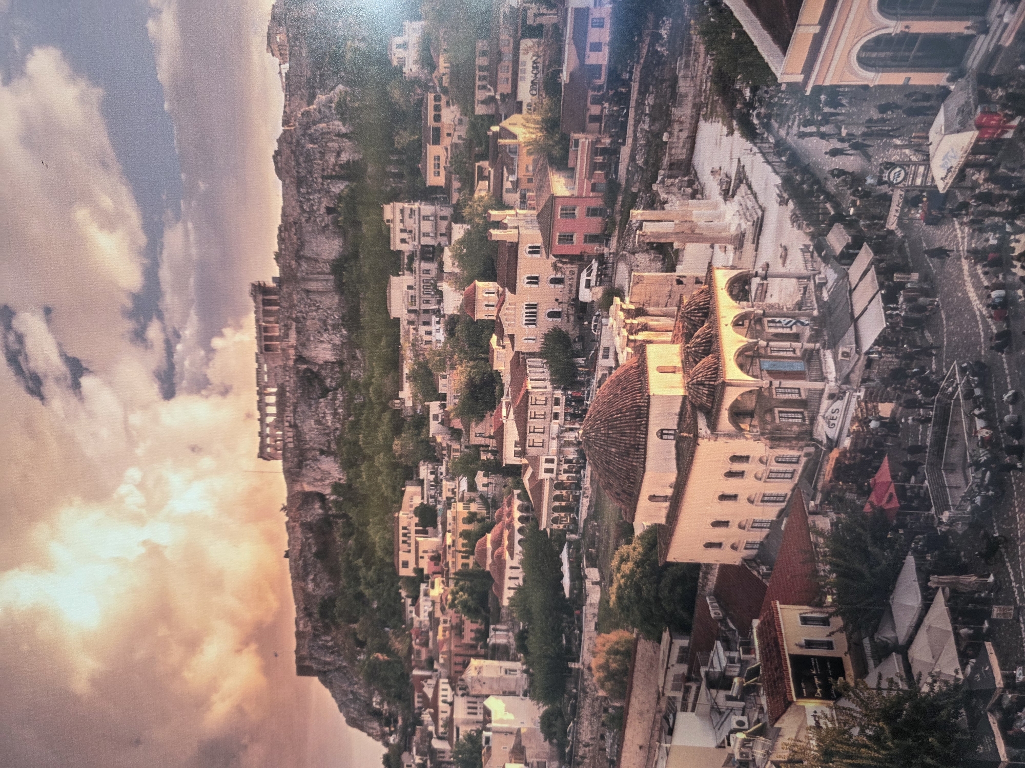 The image depicts a bustling street scene in a densely populated city, possibly in a Mediterranean or European location. The perspective is from above, looking down onto a street lined with shops, restaurants, and residential buildings. In the foreground, several people are visible, some walking and others seated at outdoor tables, suggesting a vibrant social atmosphere. Objects like umbrellas, signs, and architectural elements are scattered throughout the scene.
The time of day appears to be either late afternoon or early evening, as indicated by the warm, golden light casting long shadows and the sky showing hues of orange and pink. The weather seems pleasant, with clear skies and diffused sunlight. The architecture features a mix of styles, with classical influences visible in some of the buildings, including arches and columns. The presence of a Greek flag on one of the buildings might suggest the location is Greece.
The overall mood is lively and energetic, with a sense of community and activity. The scene is crowded with people, creating a dynamic and engaging visual. There are several instances of text visible in the image, though much of it is too small or blurry to be fully deciphered. One sign appears to read "GES," and another sign in the foreground has some Greek lettering on it. There are also some signs with English text.