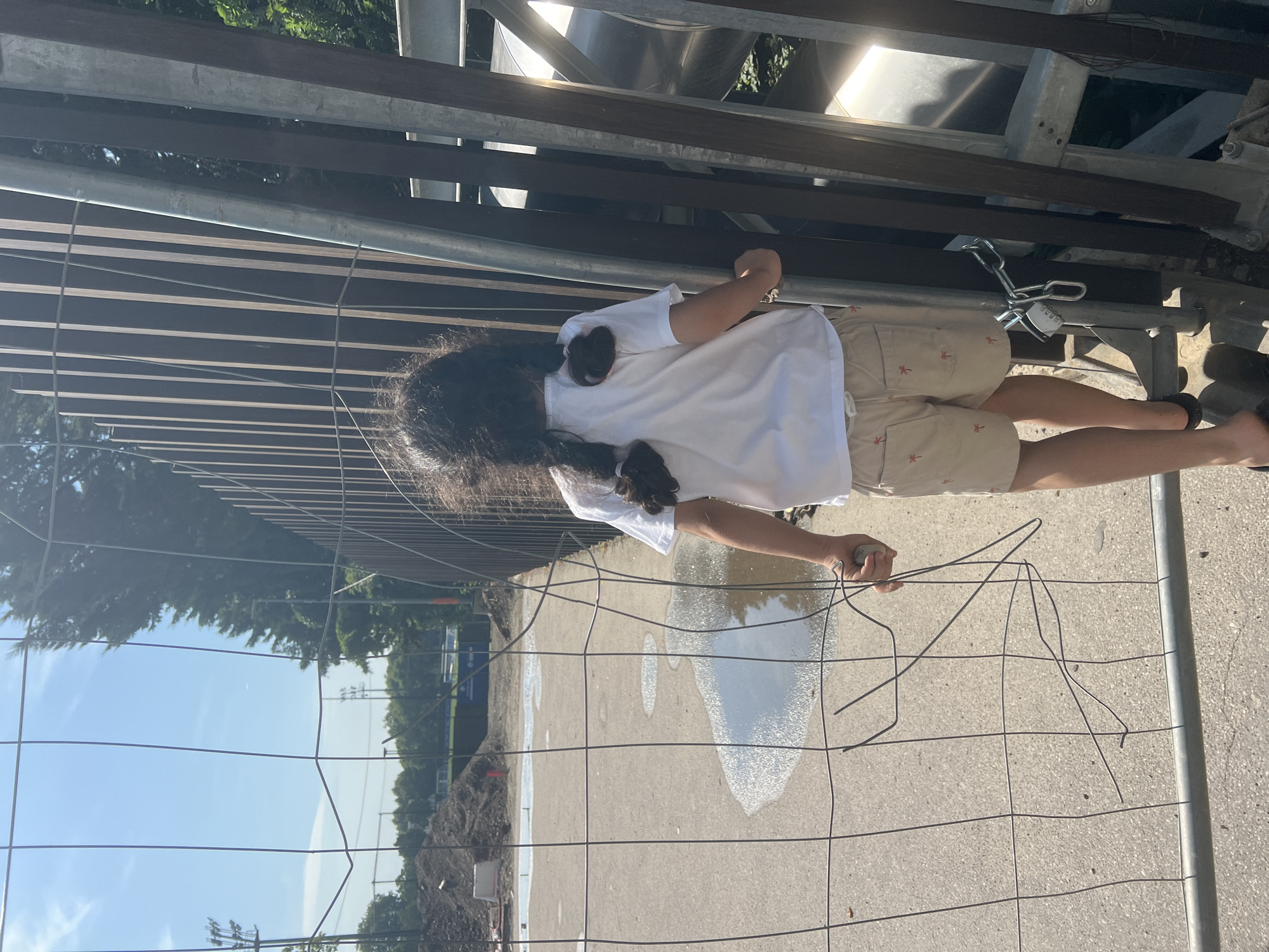 An outdoor medium shot captures a young child, facing away from the camera, standing near a metal fence in an urban environment. The child has dark, braided hair and is wearing a white t-shirt and beige shorts with a palm tree pattern. Their right hand is raised, holding a small, gray object. The fence, partially bent and damaged in the foreground, is a temporary mesh barrier. Behind the fence, there is a construction site with piles of dirt and puddles of water on a paved surface. In the background, trees and a clear blue sky are visible. The scene appears to be a sunny day.