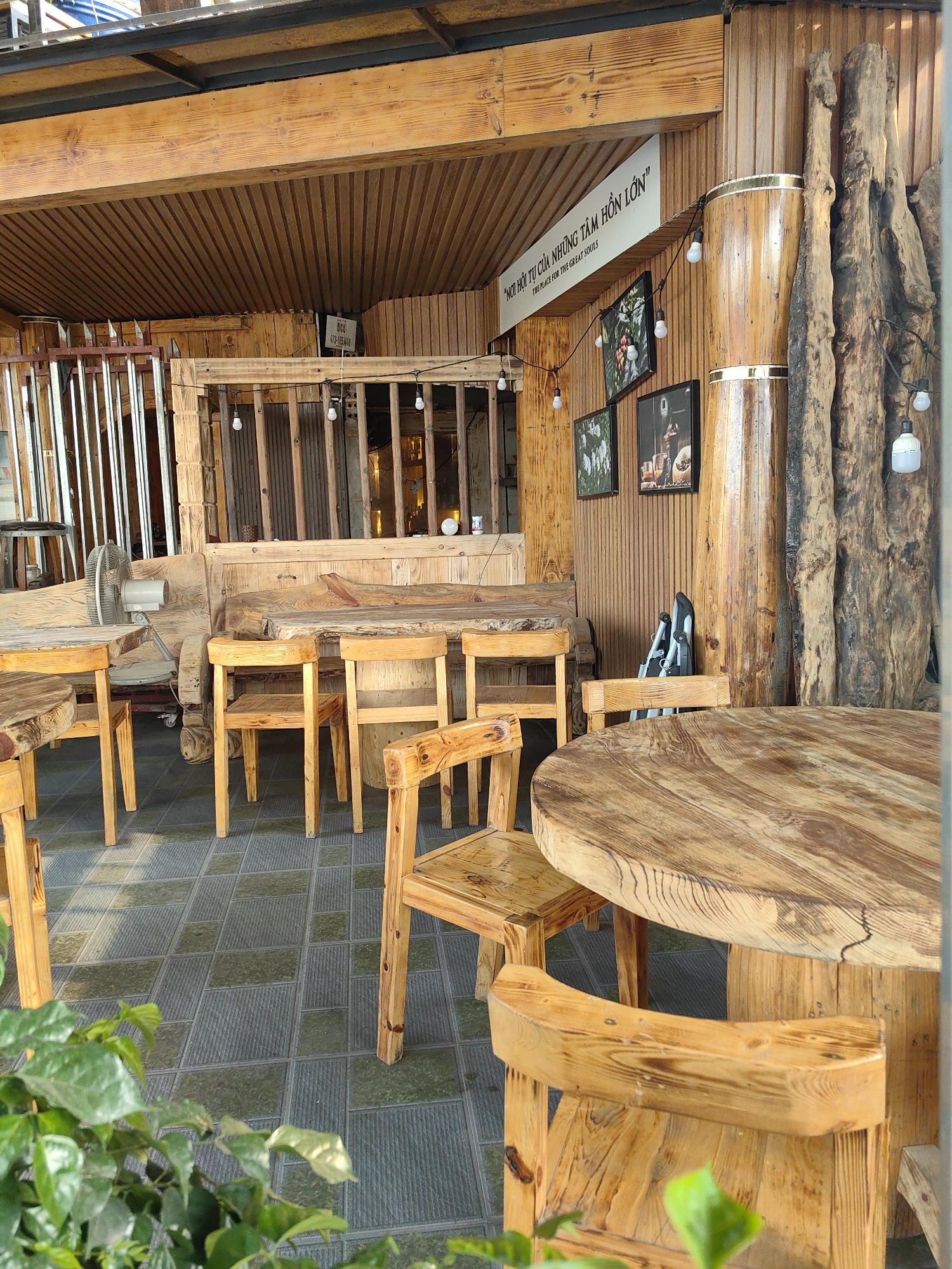 Here is a description of the image:
The photograph depicts an outdoor seating area of a wooden cafe located in Bai Chay Ward, Vietnam. The furniture consists of round and rectangular solid wood tables and matching chairs. In the background, there is a wooden bench and additional seating behind a rustic partition. The cafe's decor includes wooden pillars resembling tree trunks, string lights, framed pictures, and a sign with the words "NƠI HỘI TỤ CỦA NHỮNG TÂM HỒN LỚN" and "THE PLACE FOR THE GREAT SOULS". In the lower foreground, green plants frame the view. The floor is tiled.