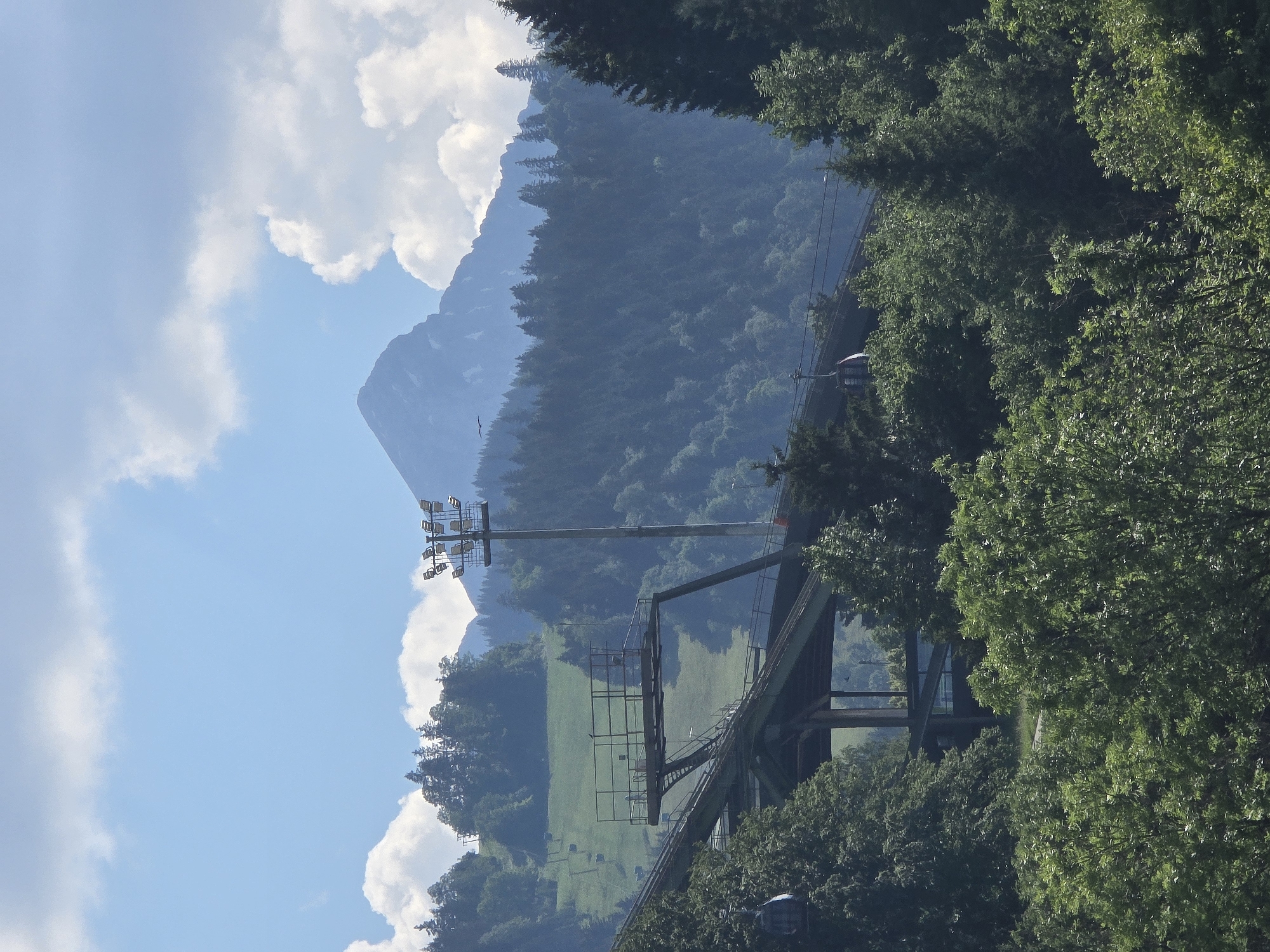 The image depicts a view of mountains and trees, with a cable car system and a large pylon with lights. The scene is set in Garmisch-Partenkirchen, Germany, likely during the daytime given the bright sky and the sunlight filtering through the trees. The weather appears to be clear with some fluffy white clouds scattered across a bright blue sky.
The primary subjects are the lush green trees that dominate the foreground and midground, partially obscuring a dramatic mountain landscape in the background. A prominent object is the cable car pylon, a tall, metallic structure with multiple spotlights attached to its top, suggesting it might be used for night events or as part of a ski resort infrastructure. A cable car cabin is visible, suspended from a cable that stretches across the frame, indicating the cable car system is operational. The cable car appears to be traveling uphill, with another cabin visible further up the track. A metal structure, possibly a viewing platform or part of the cable car station, is also visible. The mountains in the background are shrouded in a soft, hazy blue, indicating distance and depth.
The overall impression is one of a scenic outdoor location, likely a tourist or recreational area. The bright lighting and clear sky suggest fair weather, and the abundant greenery points to a warmer season. There is no visible text in the image. The scene is serene and picturesque, evoking a sense of natural beauty and outdoor adventure.