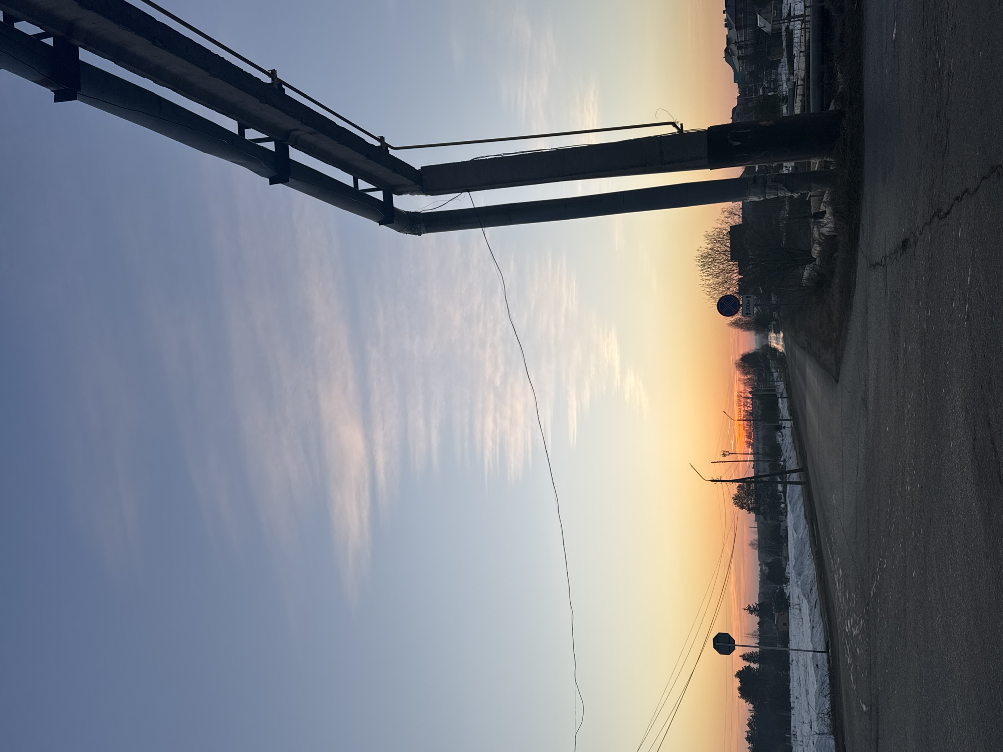This is a photograph taken in Прикамский, Russia. The image is oriented vertically and shows an urban scene during a sunrise or sunset. On the left side of the image, there are gray metallic pipes supported by black steel beams and brackets. The sky is visible behind the pipes, displaying a gradient of blue transitioning into yellow and orange near the horizon. The clouds are faint and elongated. On the right, there is a cracked asphalt road that runs along the side of the image. Alongside the road are trees and shrubs. Street signs and utility poles are also visible along the road, indicating the presence of infrastructure. Buildings are visible at a distance in the horizon. The ground has a layer of snow covering most of the landscape.