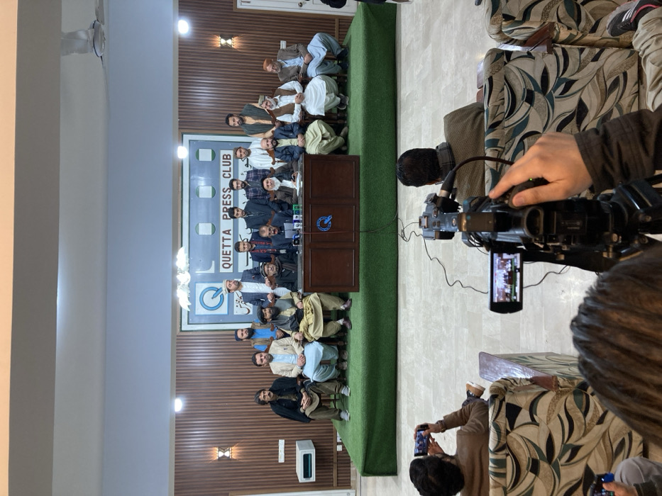 An indoor scene depicts a press conference taking place at the Quetta Press Club in Quetta City Tehsil, Pakistan. Eighteen adult males are positioned on a raised platform, approximately 30 cm high, covered in green artificial turf. Seven men are seated behind a dark wooden table equipped with multiple microphones, with one central figure speaking. Eleven other men are seated or standing behind them. Most individuals wear traditional Pakistani clothing, including shalwar kameez and waistcoats, with some wearing traditional headwear. In the background, a large white sign with a light blue border displays "QUETTA PRESS CLUB" in capital letters, along with a 'Q' logo and Urdu script. The wall behind the platform is clad in dark brown vertical paneling, featuring two sconce lights and a white wall-mounted air conditioning unit on the left. The ceiling is white with recessed lighting. In the foreground, from a lower perspective, at least three media personnel are visible from behind, seated in patterned armchairs. One individual on the left records the event with a horizontally-held smartphone. Another, centrally positioned, operates a black professional video camera on a tripod, its flip-out screen displaying the view of the platform. The floor in the audience area is light-colored polished tile.