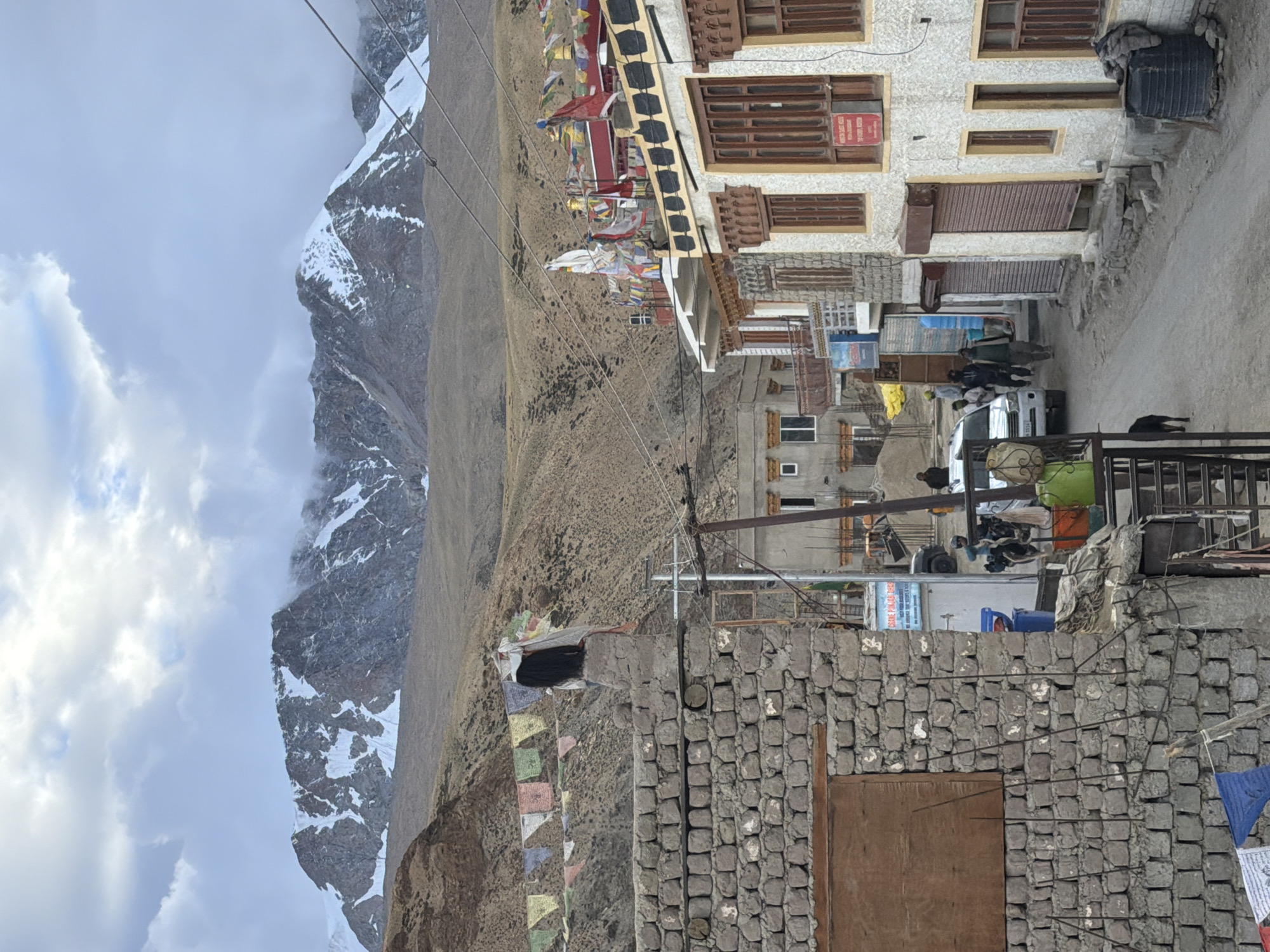 In Karzok, India, this image captures a street scene with a prominent stone wall on the left and buildings lining the right side of a dirt road. In the background, snow-capped mountains rise under a cloudy sky, with prayer flags strung across the landscape. A white SUV is parked on the road, and several people are walking or standing nearby. The buildings exhibit traditional architecture with wooden window frames and decorative elements. A dark-colored dog is visible on the lower left side of the image. The overall atmosphere suggests a remote, mountainous village with a blend of human activity and natural grandeur.