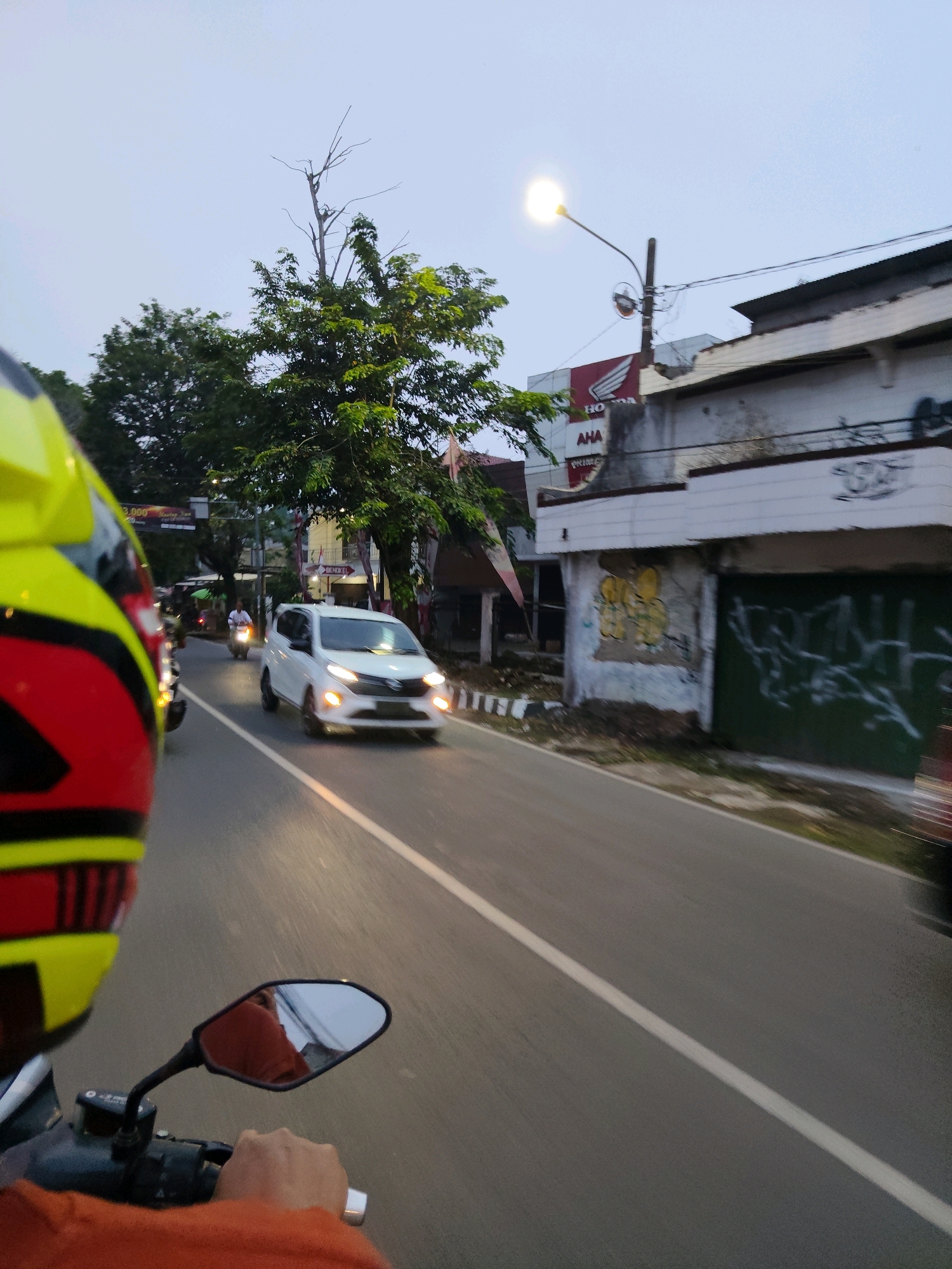 The image captures a street scene in Kaduagung Timur, Indonesia, from the perspective of someone riding a motorcycle. In the foreground, the left side of a yellow, red, and black helmet is visible, along with the rider's orange shirt and hand gripping the handlebar. The motorcycle is in motion on an asphalt road marked with a white dividing line. Ahead, a white hatchback car is traveling in the same direction, its headlights illuminated. To the right of the road, there is a building with a white facade, featuring graffiti and boarded-up windows. A street light with a bright bulb is positioned above the building. Trees with green foliage line the left side of the road, interspersed with other buildings and some traffic lights in the distance. The sky is overcast, suggesting it might be dusk or dawn. The overall impression is one of everyday traffic and urban infrastructure in a town.