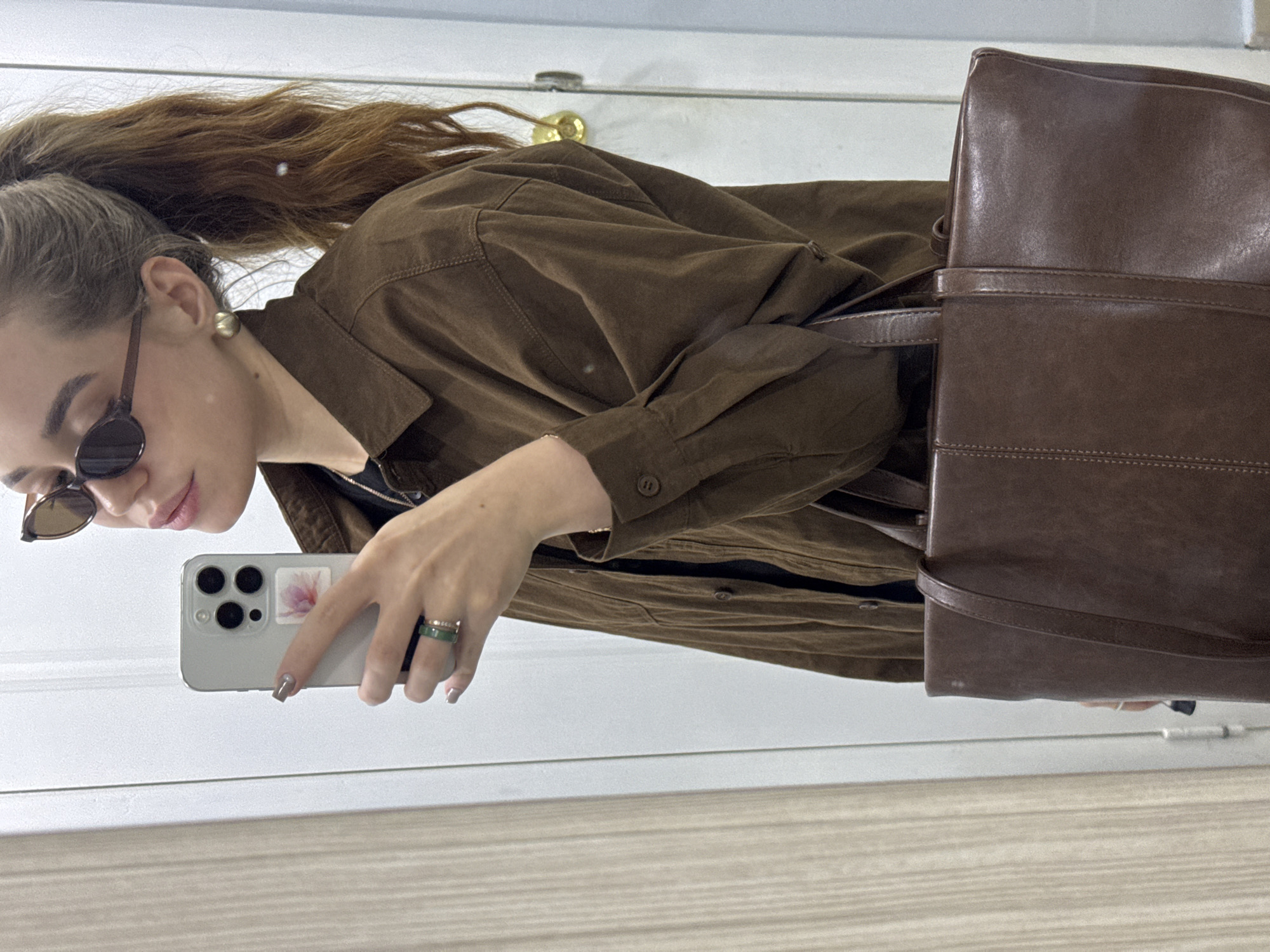 A woman is taking a selfie in a mirror. She has brown hair in a high ponytail, sunglasses, and a brown button-down shirt. She is holding a smartphone with a floral sticker on the back. She is wearing rings on her fingers. A brown leather bag is partially visible. The background includes a white door. There is no indication that this image was taken in Chaikovsky, Russia.