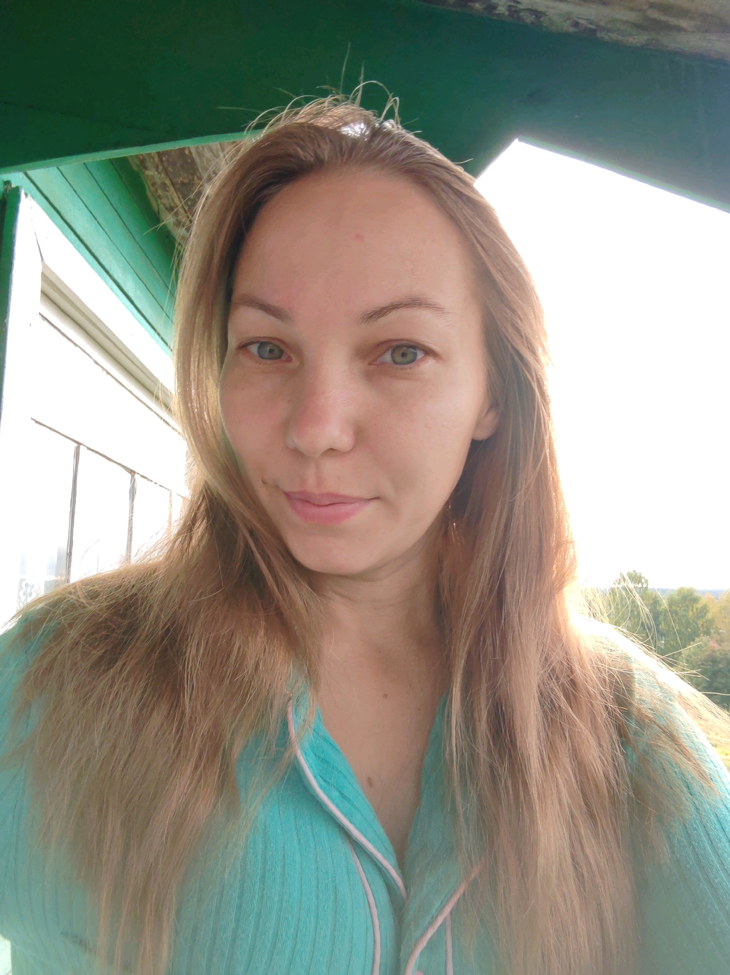 The image is a selfie taken by a Caucasian woman with long, light brown hair. She is wearing a teal-colored ribbed top with pink piping. Her eyes are blue-green, and she has a mole on her right cheek. She is outdoors, with a green wooden structure above her and a window with vertical panes to her left. The background shows trees and a bright sky. The woman is looking directly at the camera with a slight smile. The location is identified as Chukhlem, Russia, though this context is not visually evident in the image itself.
