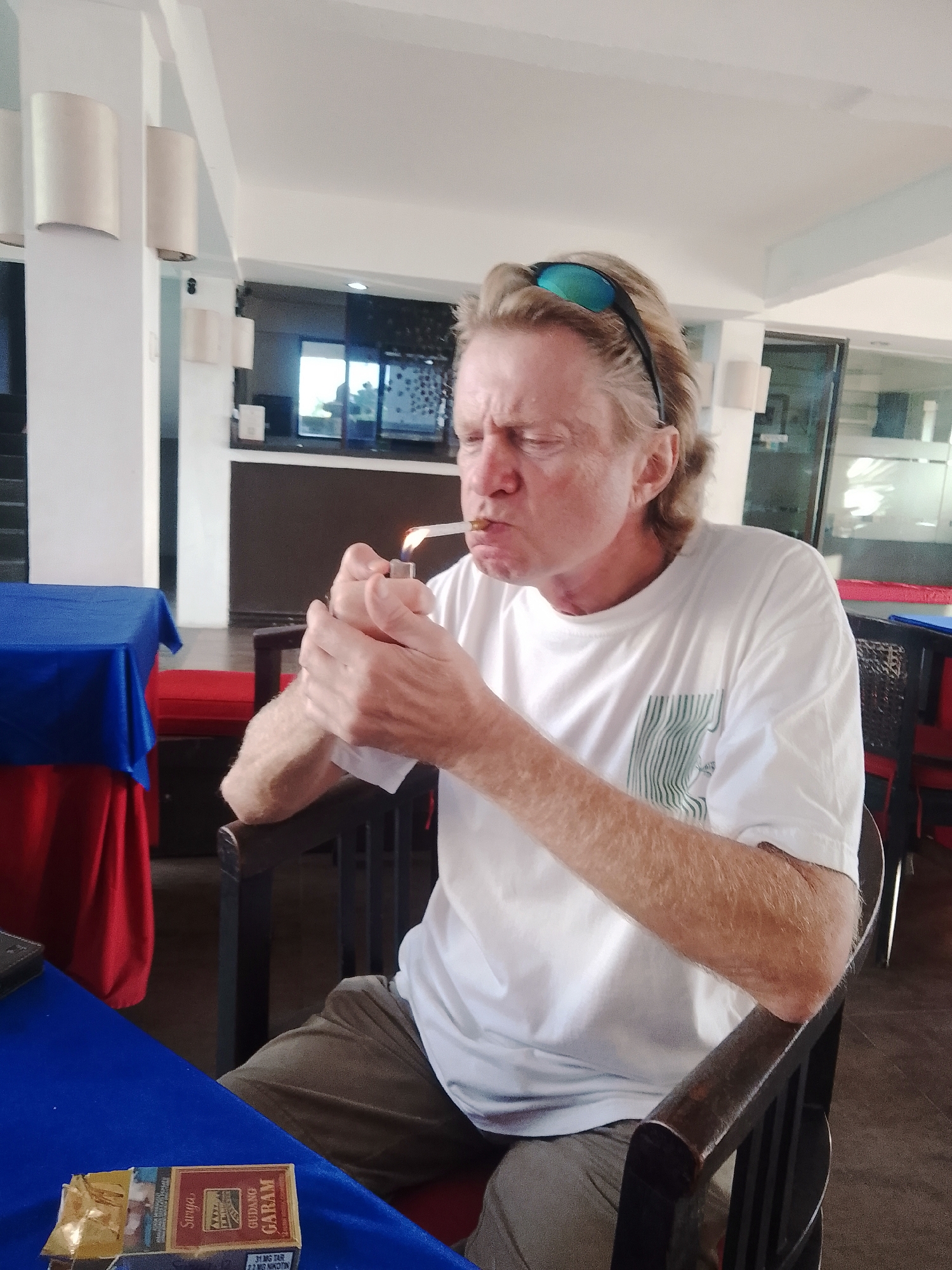 A middle-aged Caucasian male with blonde hair and sunglasses perched on his head is lighting a cigarette with a lighter. He is wearing a white t-shirt and brown pants. The man is sitting at a table covered with a blue tablecloth, and a pack of Gudang Garam cigarettes is visible on the table. The background suggests an indoor establishment with decorative lamps and seating arrangements. The setting appears to be a restaurant or cafe in Denpasar, Indonesia, given the presence of Gudang Garam cigarettes, a popular Indonesian brand.