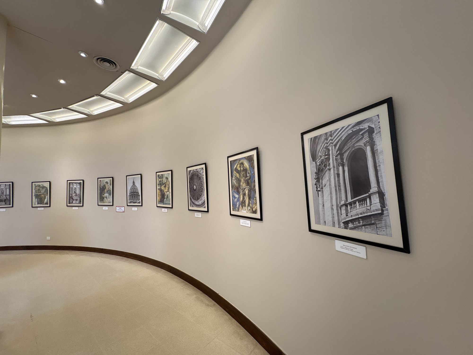 The image depicts an interior view of a well-lit, unoccupied art gallery or exhibition space within San Carlos de Bariloche, Argentina. The scene is dominated by a light-colored, gently curved wall displaying a series of nine framed artworks, extending from the foreground on the right into the background on the left. Each artwork, varying in subject from classical architectural elements like a balcony and a dome to intricate religious or historical figures and designs, is presented in a dark frame with white matting and has a small white descriptive plaque affixed to the wall below it. For instance, the prominent image on the far right is a black and white photograph of a classical "BALCÓN EXTERNO" (external balcony) from the "Basilica di San Pietro in Roma" (St. Peter's Basilica in Rome). The floor is a uniform light-colored surface, possibly carpet or smooth tile, with a dark, narrow trim delineating its edge along the base of the curved wall. The ceiling, matching the wall color, features a series of rectangular, recessed fluorescent light panels arranged in an arc, supplemented by several small, circular recessed spotlights and a circular air vent, providing consistent illumination to the space. No individuals are present in the scene, and no actions or interactions are depicted.