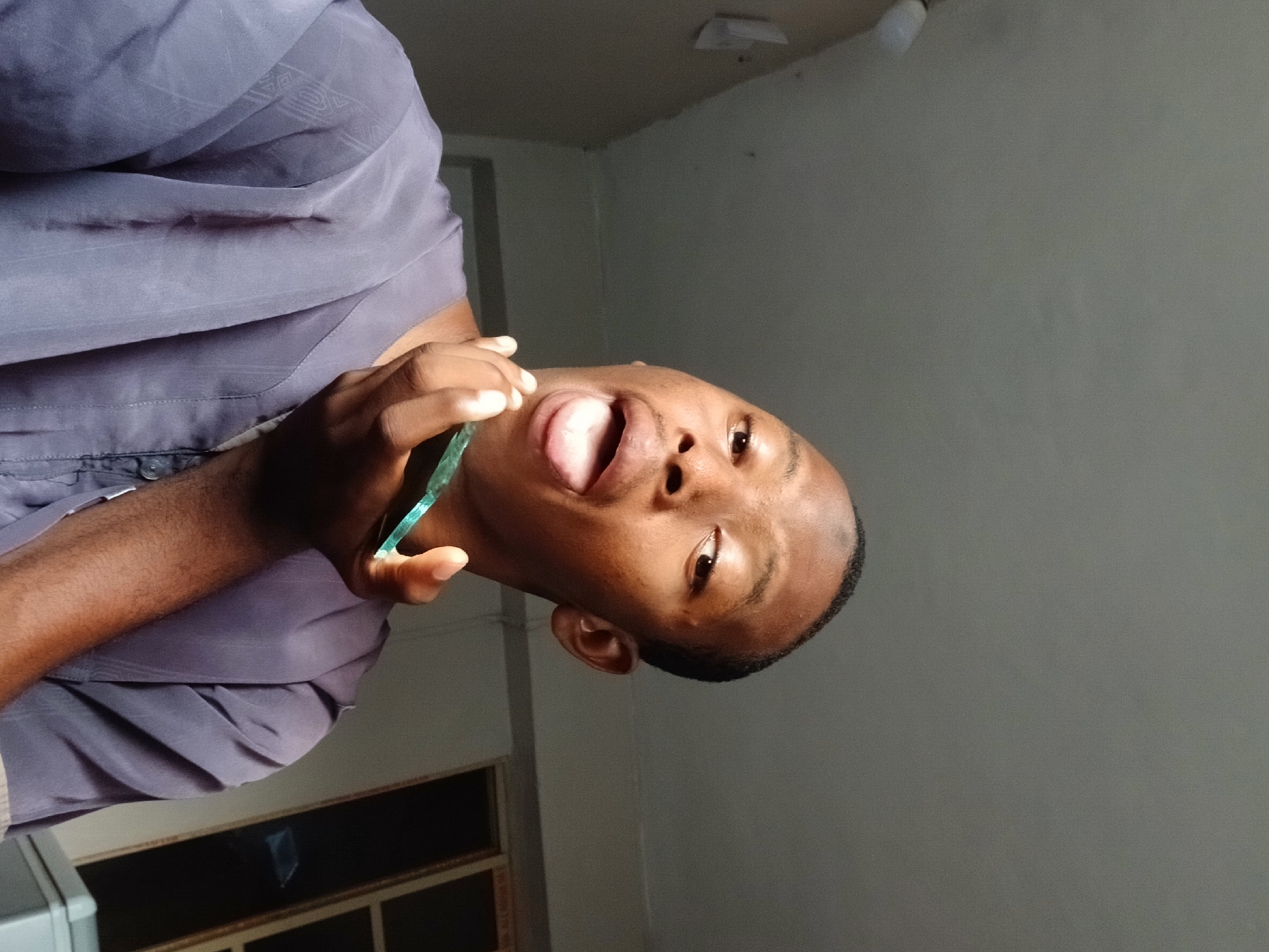 A young man with brown skin and close-cropped dark hair is holding a translucent green object to his mouth, possibly a toothbrush. He is wearing a gray shirt with a patterned design. The background appears to be an indoor setting with a gray wall and a window or cabinet. There is no definitive evidence to confirm the location is Lagos, Nigeria.
