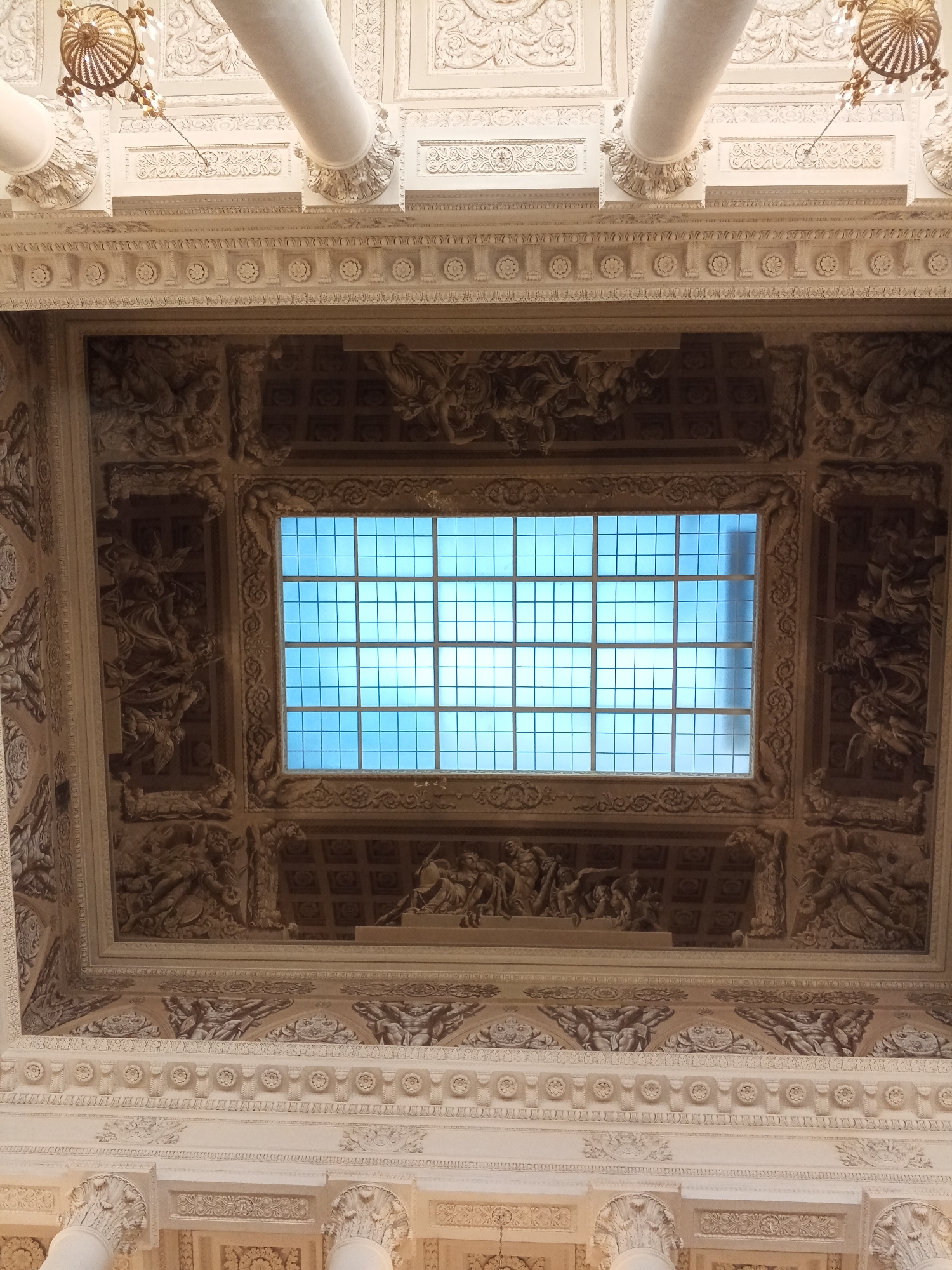 The image captures a highly ornate ceiling within a building in Saint Petersburg, Russia. The ceiling is primarily white and tan, with intricate detailing, including sculpted figures and geometric patterns. A rectangular skylight fills the center of the frame, its grid of glass panels allowing bright blue light to filter through. The surrounding ceiling sections are elaborately decorated with carvings depicting what appear to be mythological scenes. The architecture includes Corinthian-style columns and chandeliers, suggesting a grand, historic interior, possibly a museum or palace.
