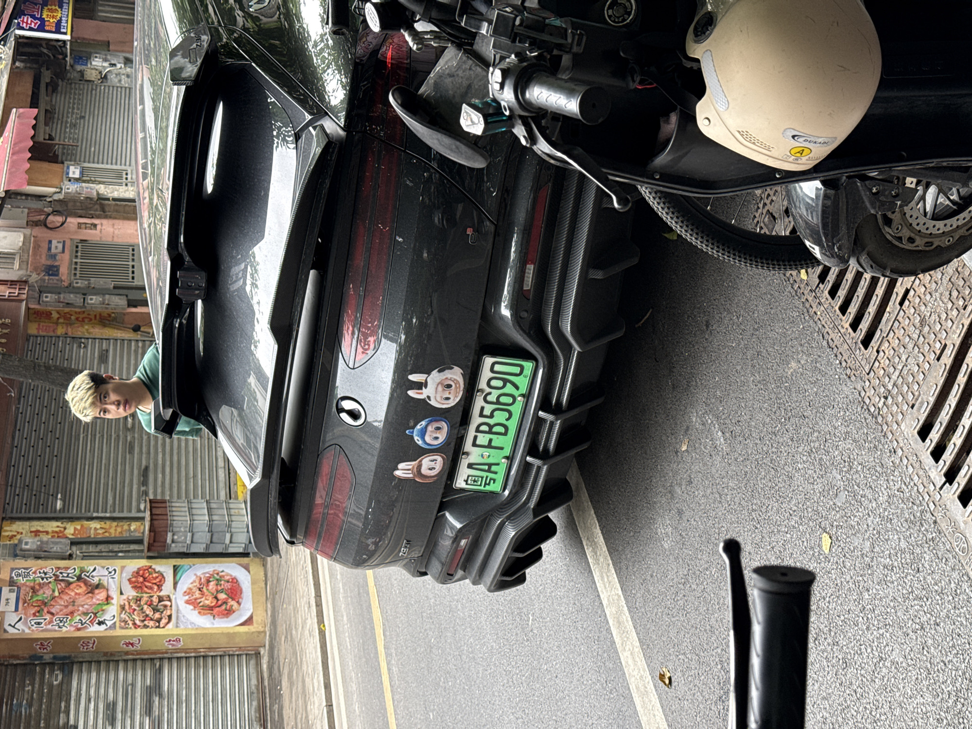 The image shows the rear end of a black car parked on a street. The car has a green license plate reading "A·FB5690" and cartoon stickers above it. Next to the car, on the right side of the image, there is a motorcycle, and a helmet is visible on top of it. A man is standing near the back of the car, looking towards the camera. A restaurant sign featuring food dishes hangs on a shuttered storefront. The street is paved and has a metal grate on the right side. The scene appears to be on a narrow street lined with shops in the Liwan District of China, suggested by the license plate prefix, which is associated with Guangdong province.