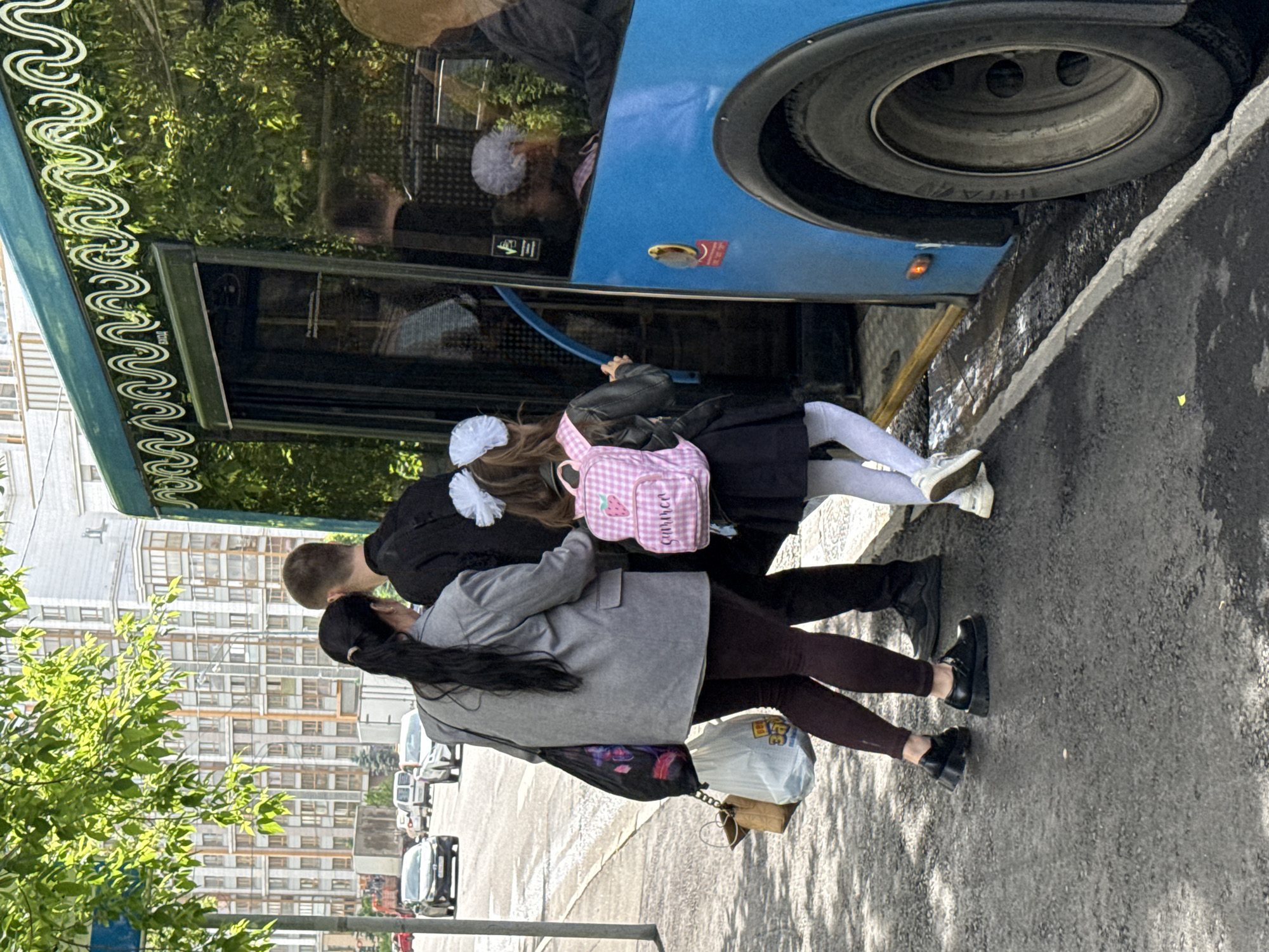 The photograph captures a moment in an urban environment, specifically a street in Moscow, Russia. A blue bus dominates the frame on the right side, positioned close to the sidewalk. A group of people are near the bus, seemingly engaged in boarding or alighting. A young girl with white bows in her hair and a pink backpack is stepping onto the bus. An adult male is nearby, possibly accompanying the child. Another adult woman, wearing a grey jacket and carrying bags, stands close to them on the sidewalk.
The street scene includes parked cars, visible trees, and a tall building in the background. The overall lighting suggests a bright, possibly sunny, day. The scene conveys a sense of everyday life and activity in a bustling urban setting.
