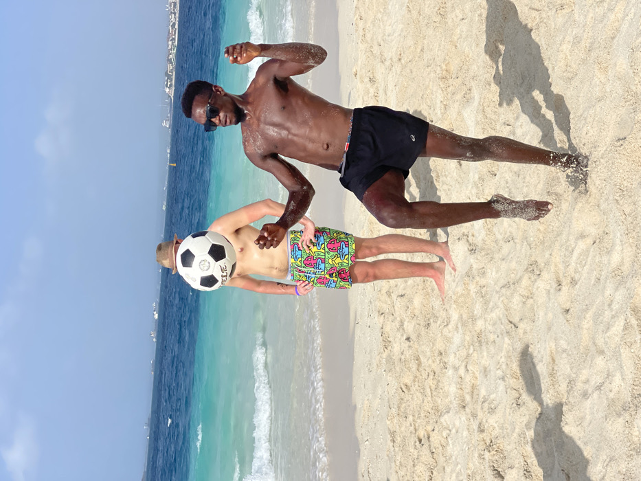 The image displays a beach scene in Zanzibar, Tanzania. Two males are present on a sandy beach under a clear, light blue sky.
In the foreground, a male with dark skin stands shirtless, wearing black swim shorts and dark sunglasses. He is captured in a dynamic pose, balanced on his right foot with his left leg lifted, knee bent, and arms flexed with hands slightly open. His skin appears wet, and grains of sand adhere to his lower legs and feet.
Positioned behind him, a male with lighter skin stands stationary, shirtless and barefoot. He wears a straw hat and multicolored swim shorts featuring a patterned design. A black and white soccer ball, marked with the word "SYELTE," is held or positioned directly in front of his face, completely obscuring it.
The immediate background consists of clear ocean water, transitioning from a light turquoise near the shore, where small waves are breaking, to a deeper blue further out. The distant horizon reveals a city skyline, identifiable as Zanzibar City, with several boats and ships visible on the water.