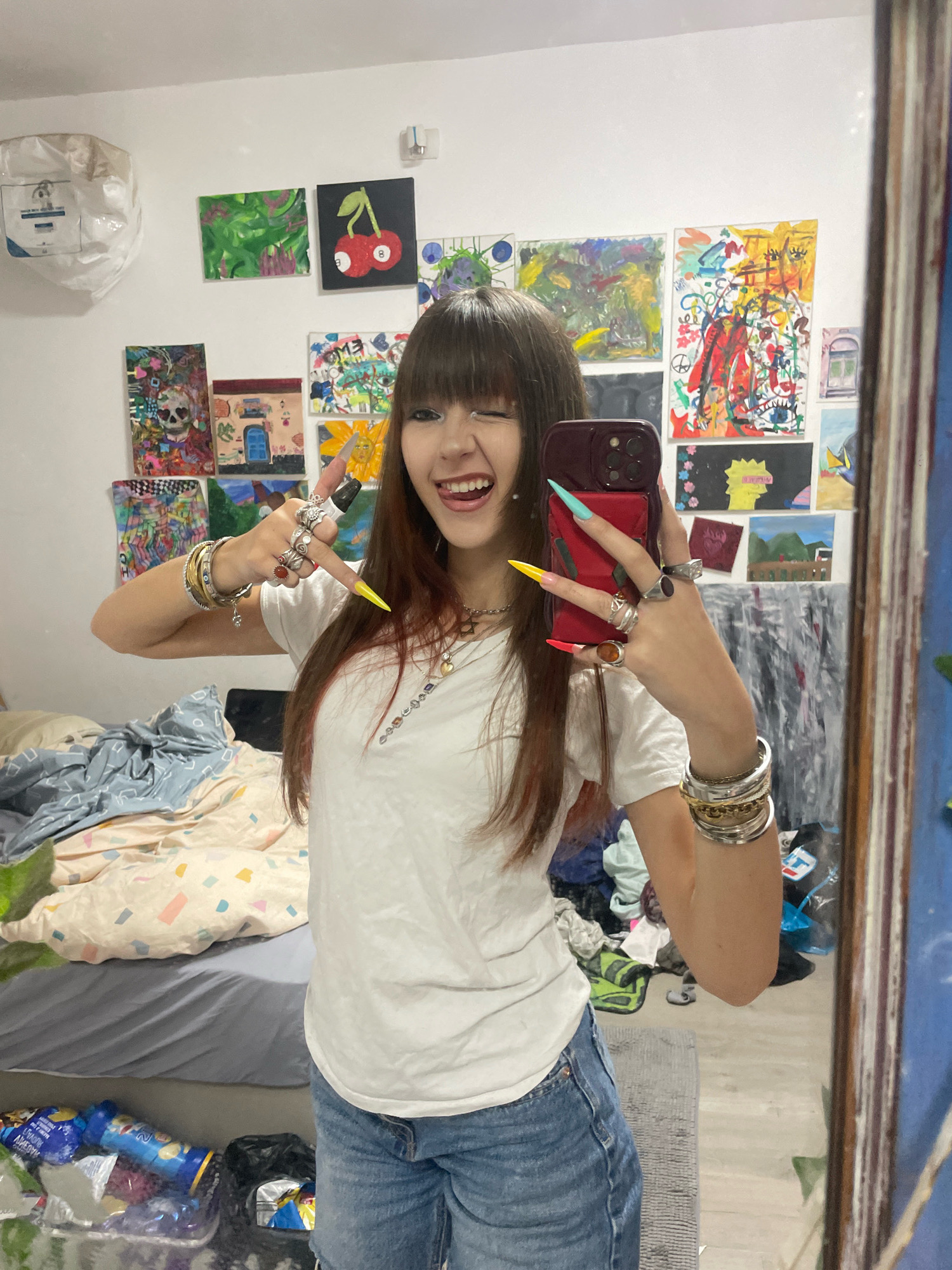 A young woman with long, dark hair and bangs is taking a mirror selfie in a room in Raanana, Israel. She is wearing a white t-shirt and blue jeans. She is smiling and sticking her tongue out, with her left hand in a peace sign and her right hand holding a phone. Her nails are long and painted in various bright colors. She is wearing several rings and bracelets on her arms. The room is decorated with many colorful paintings on the walls. A bed with rumpled bedding is visible in the background, along with some scattered items on the floor. A trash bag is also visible on the floor. The overall impression is of a vibrant and personal space.